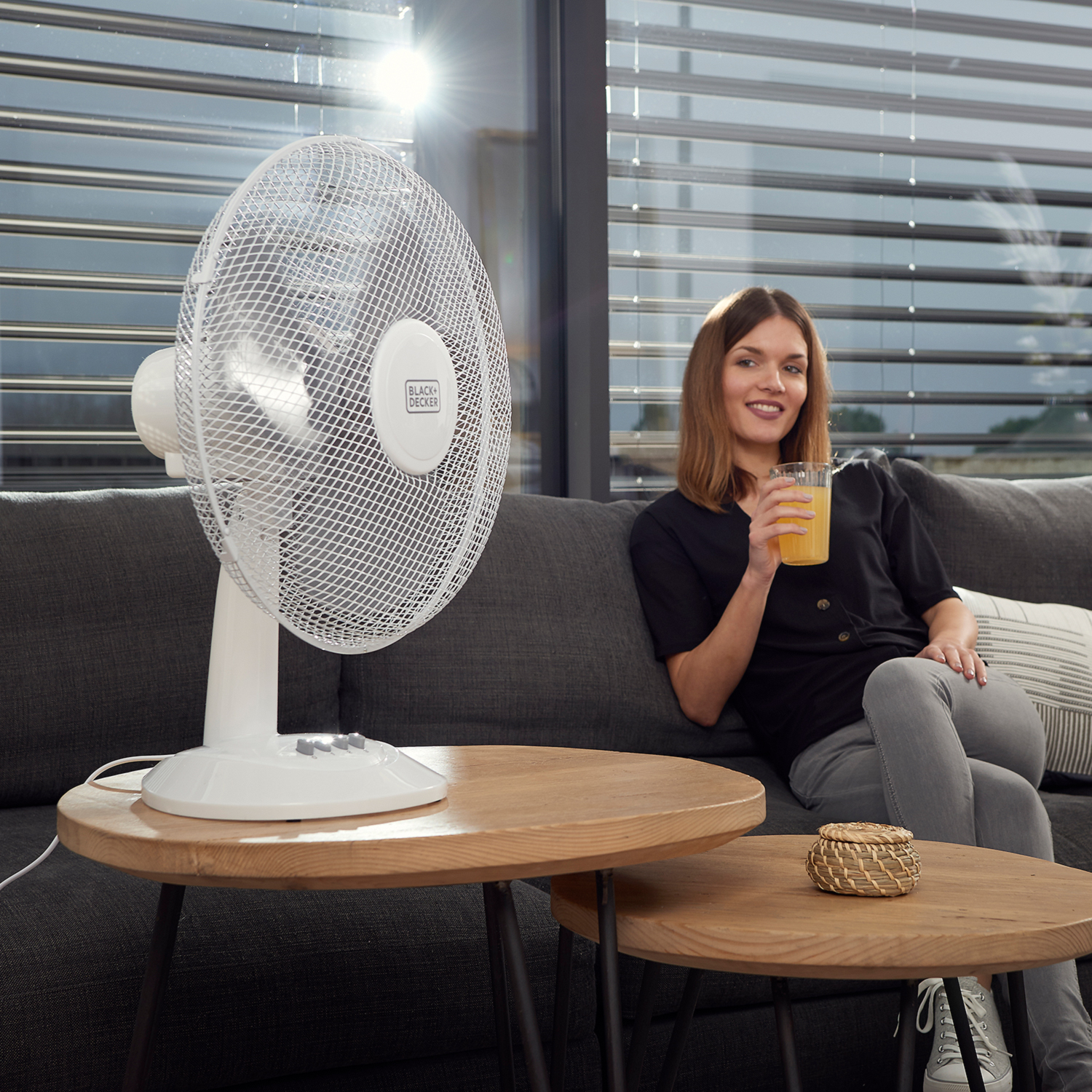 Una donna è seduta su un divano grigio con un ventilatore e un tavolo. Tiene una bevanda.