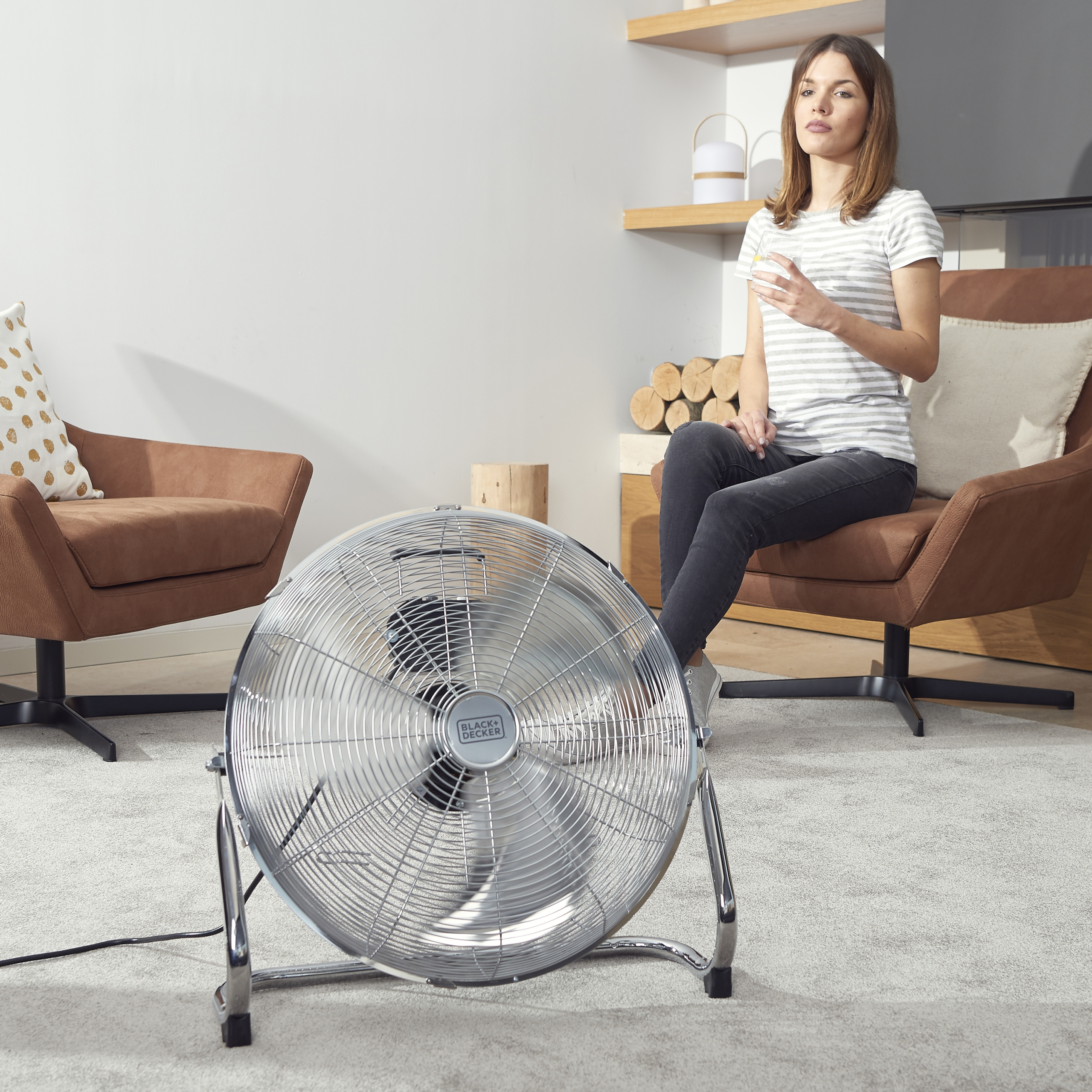 Donna seduta vicino a un ventilatore argentato. Sedie marroni, parete bianca e mensola con tronchi.