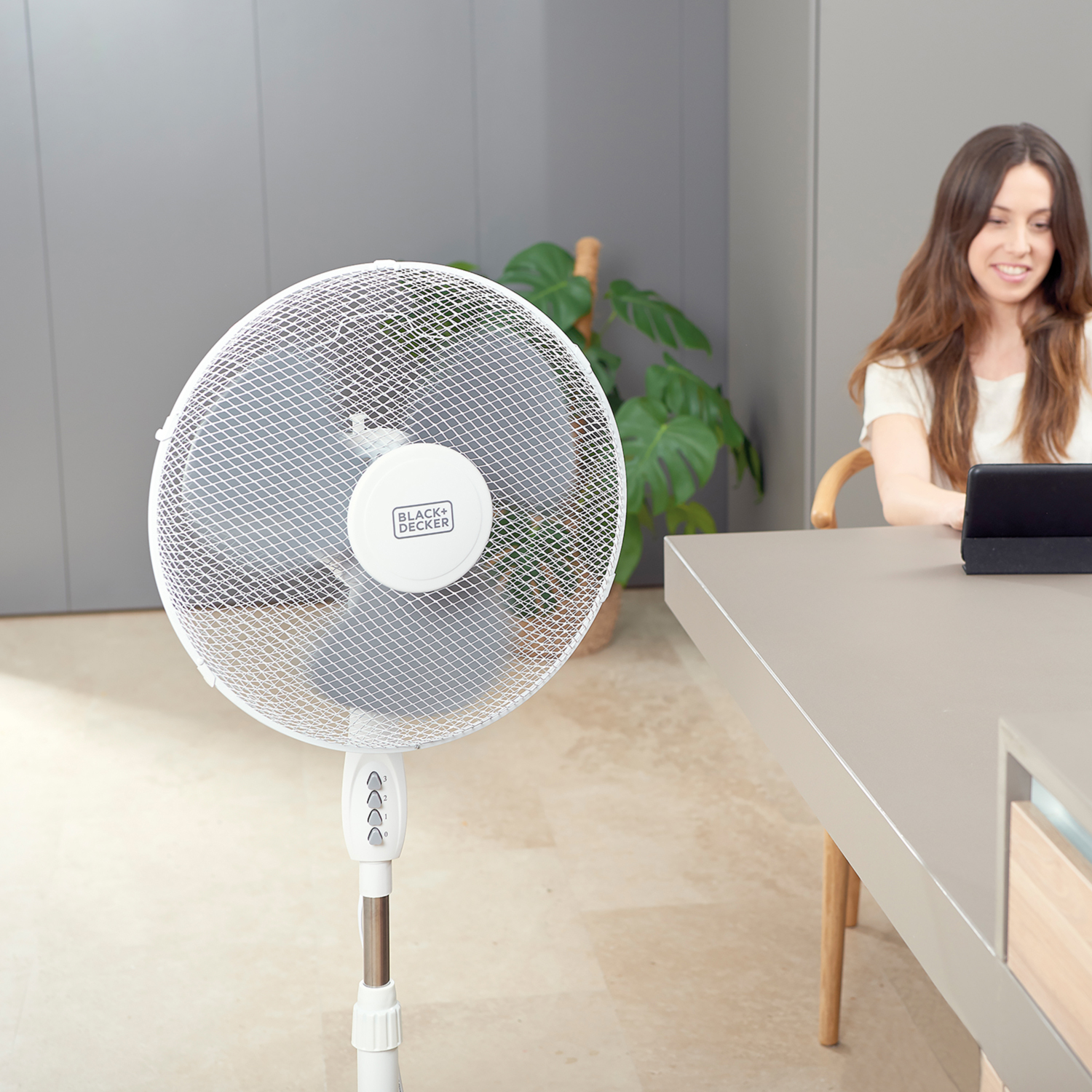 Ventilatore a piantana bianco con griglia, persona che lavora su tablet e pianta in una stanza.