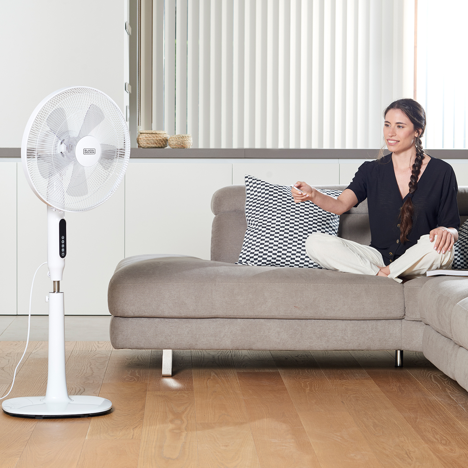 Una donna su un divano usa un telecomando per controllare un ventilatore bianco. Il ventilatore è su un supporto.
