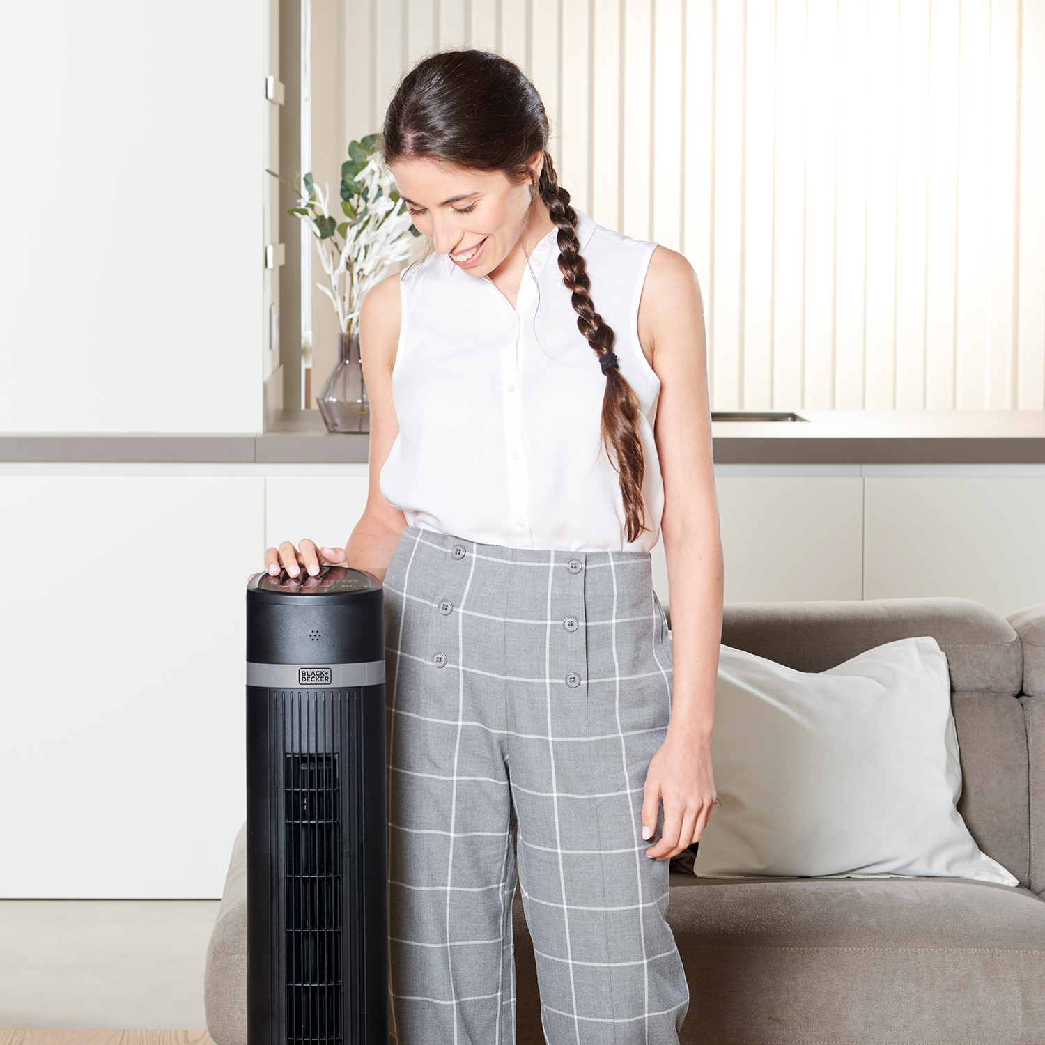 Mujer cerca de un ventilador de torre negro. Lleva camisa blanca y pantalón gris. Ambiente interior.