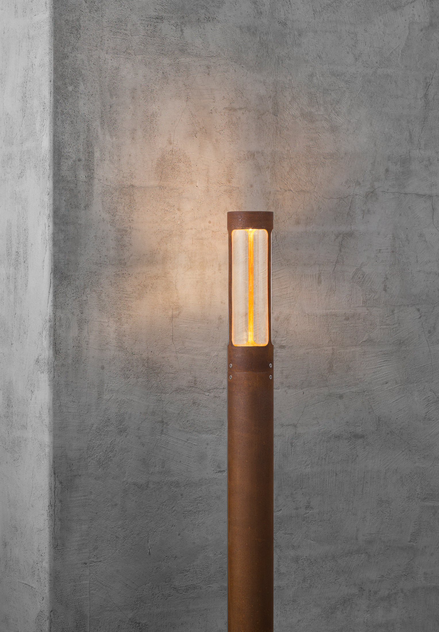 Lampada da esterno alta, marrone e illuminata contro un muro strutturato.