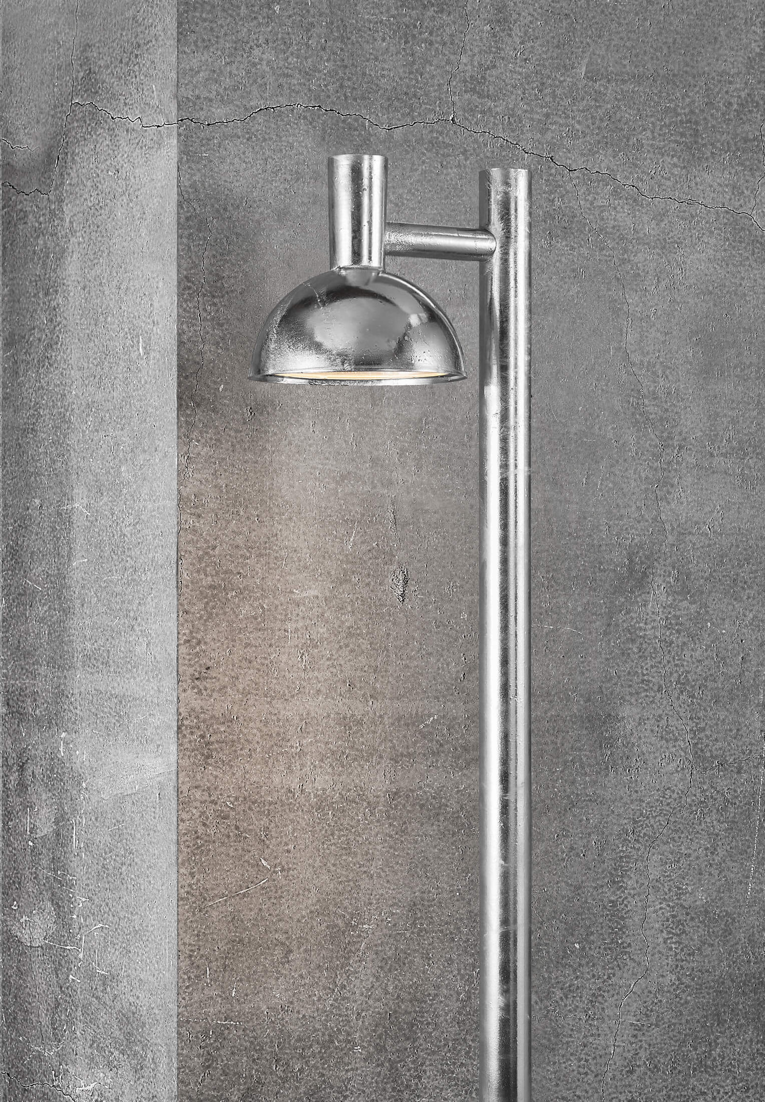 Eine silberne Außenlampe. Sie hat einen geschwungenen Schirm und eine lange vertikale Stange. Der Hintergrund ist eine Betonwand.