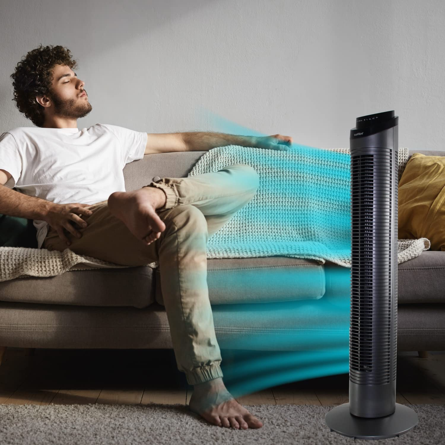 Hombre relajándose con un ventilador soplando aire. El ventilador es negro, y el hombre está en un sofá.