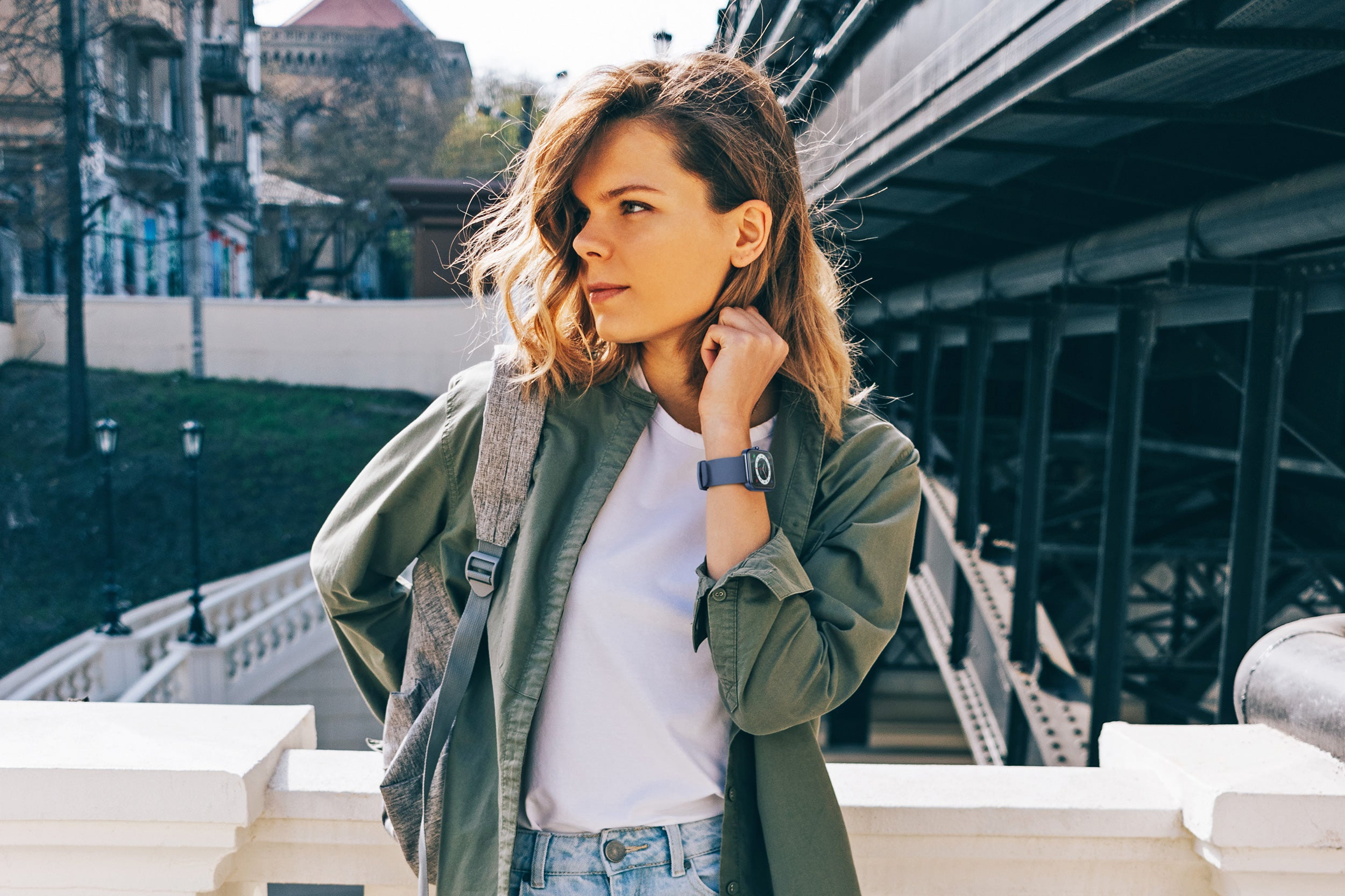Mujer con chaqueta verde, camisa blanca, vaqueros, mirando hacia otro lado. Está cerca de una barandilla blanca y un puente.