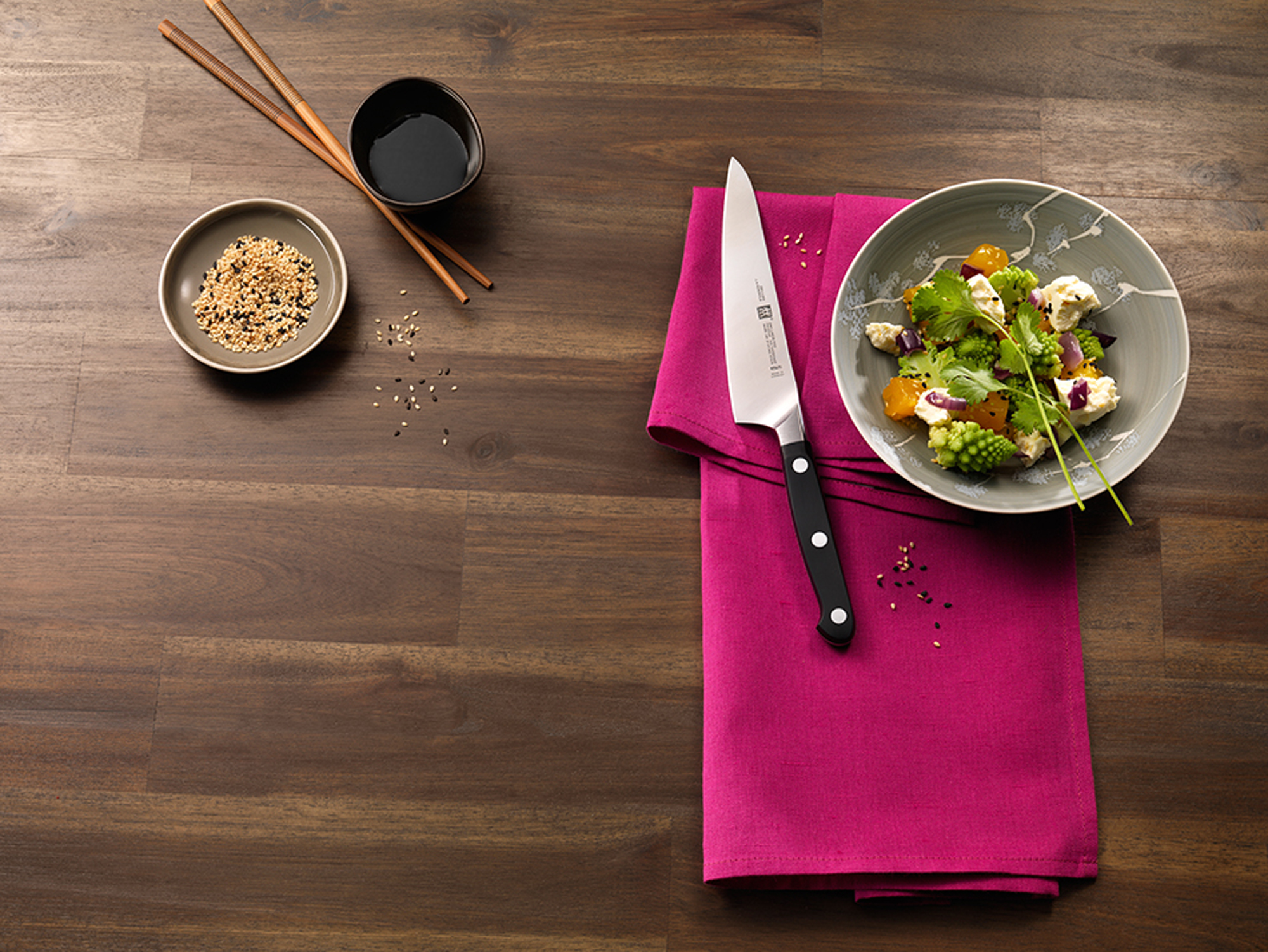 Ustawienie stołu z jedzeniem. Nóż leży na różowej serwetce obok miski z sałatką. Pałeczki i nasiona sezamu są w pobliżu.
