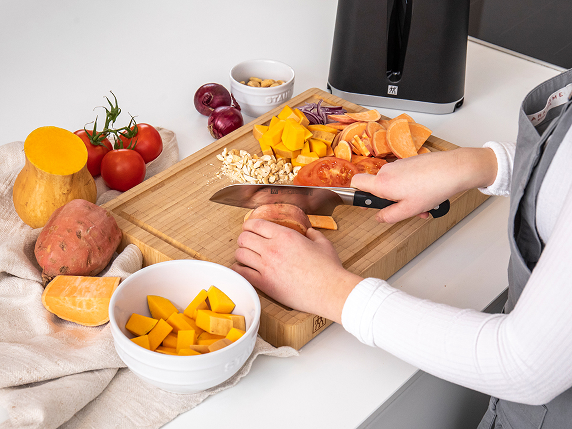 Osoba kroi warzywa na drewnianej desce. Pomidory, dynia, bataty i cebula znajdują się na desce.