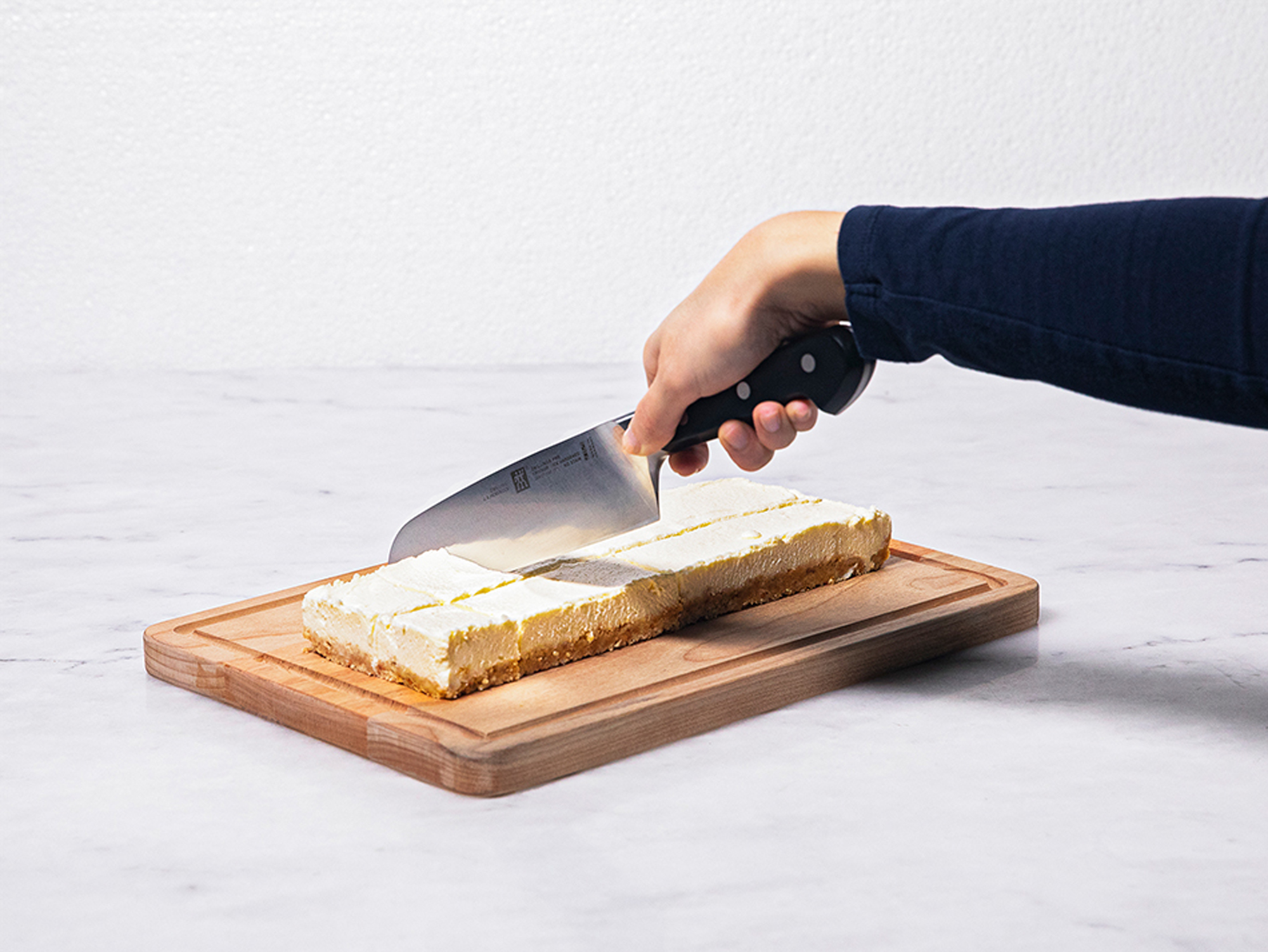 Osoba kroi sernik na drewnianej desce. Nóż przecina deser, który leży na marmurowej powierzchni.