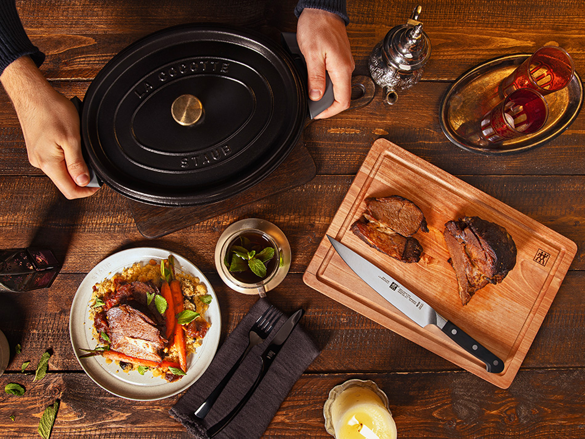 Stół do posiłku. Zawiera czarny garnek, talerz z jedzeniem, deskę do krojenia z mięsem i napoje.