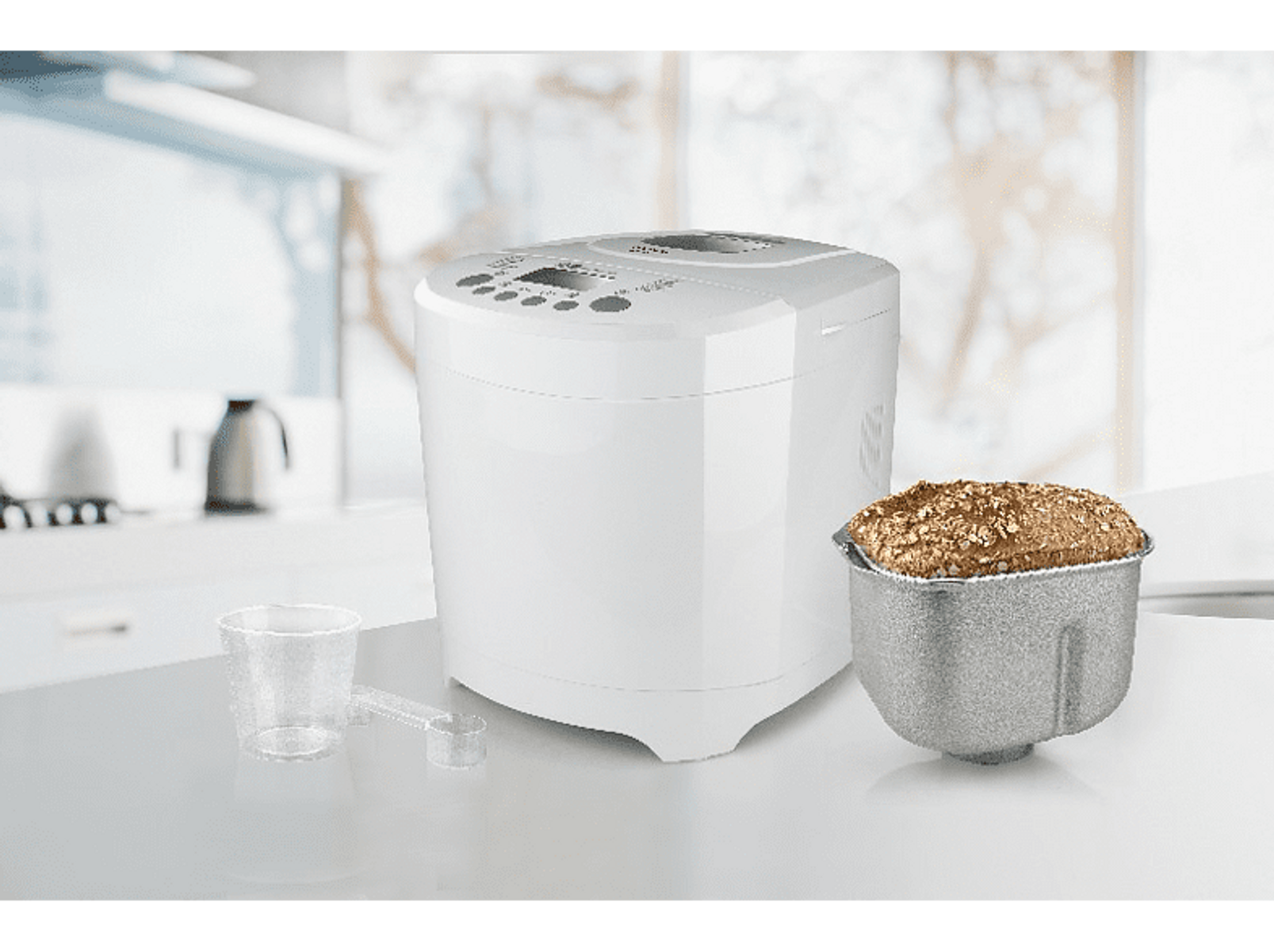 Una panificadora blanca con pan y tazas medidoras en una encimera blanca.