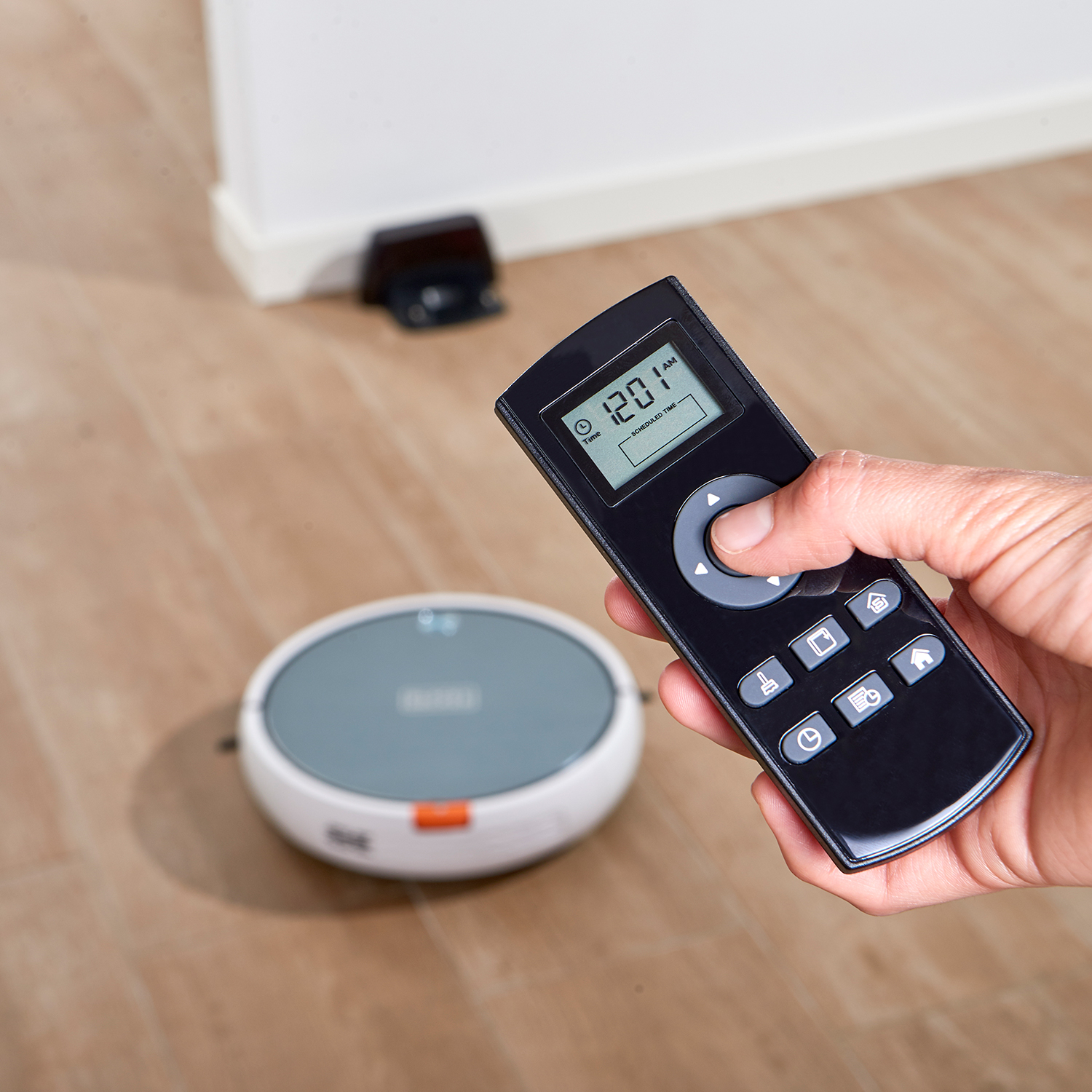 Ręka trzymająca czarny pilot wskazujący na robota sprzątającego na drewnianej podłodze.