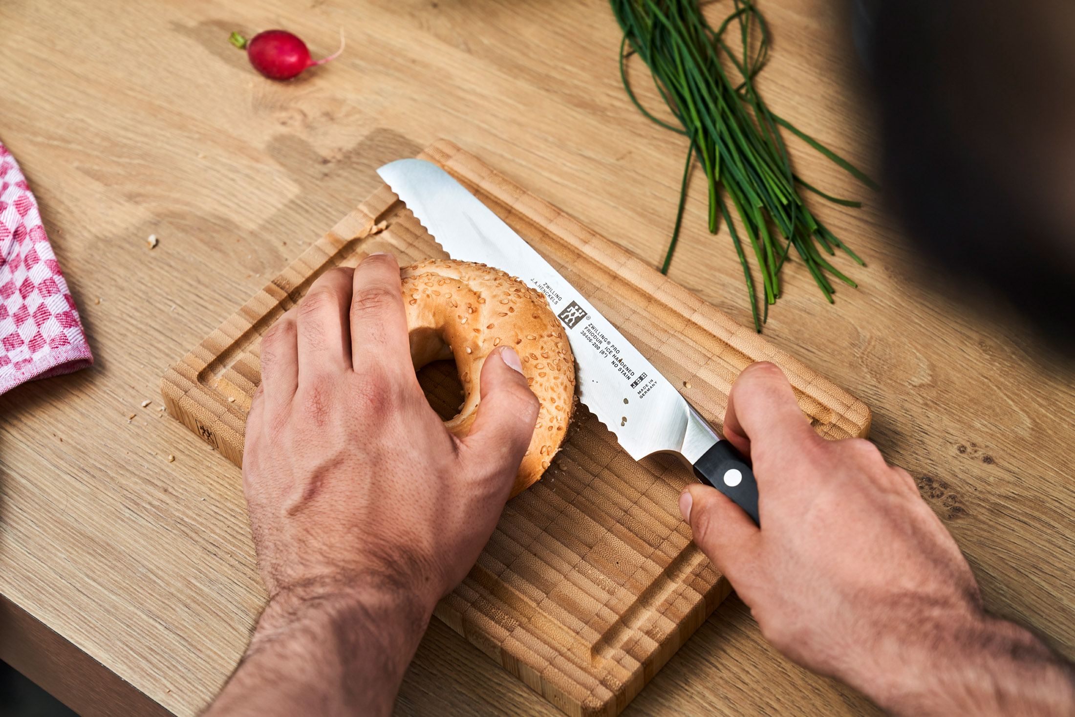 Osoba kroi bajgiel na drewnianej desce do krojenia nożem. Szczypiorek i rzodkiewka są w pobliżu.