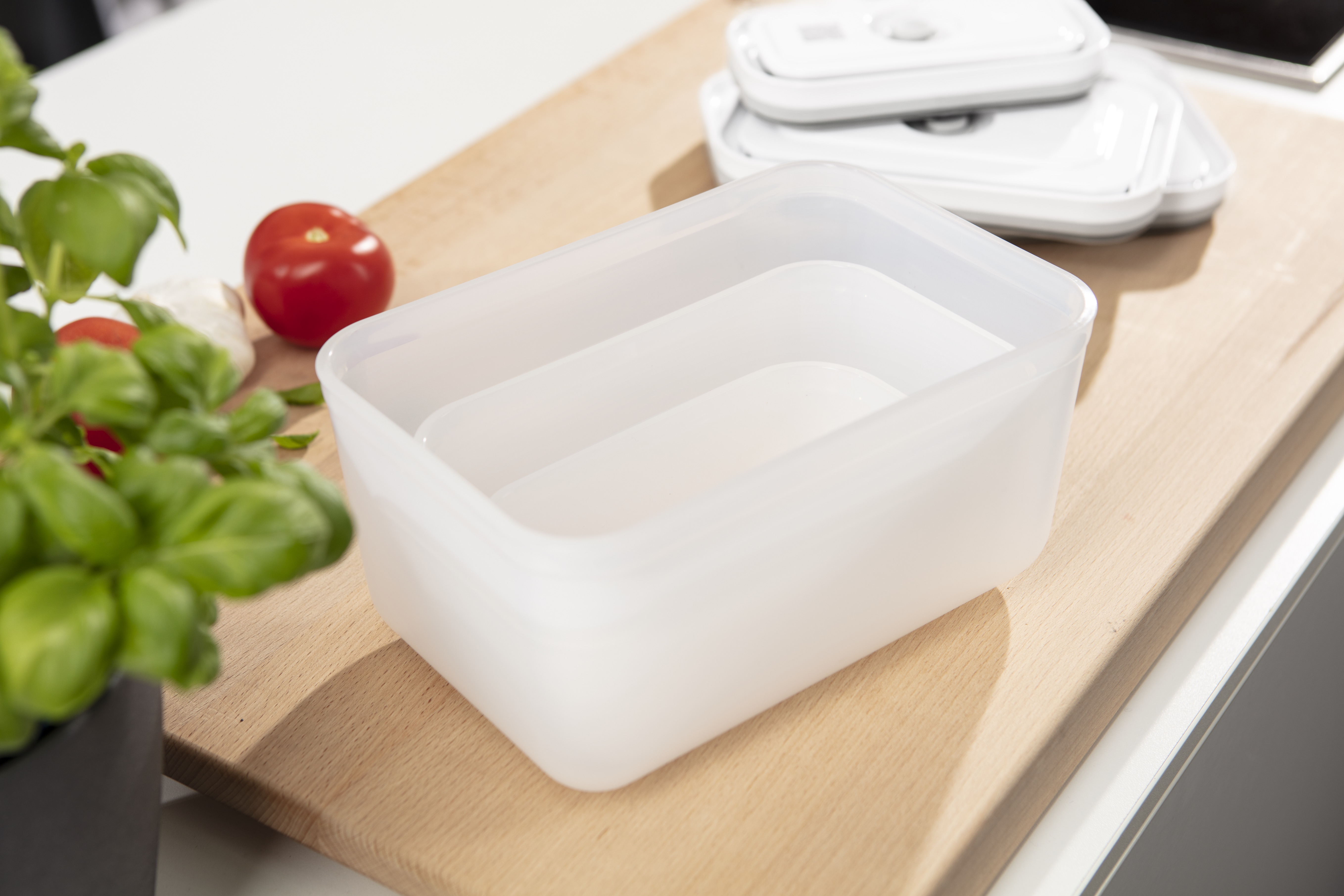 Un recipiente de plástico transparente en una tabla de cortar de madera con tomates y albahaca.