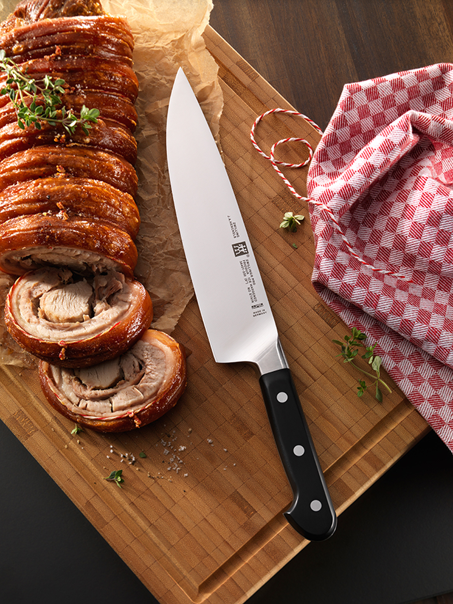 Nóż spoczywa na drewnianej desce obok porchetty. Widoczny jest również czerwono-biały sznurek z ziołami.