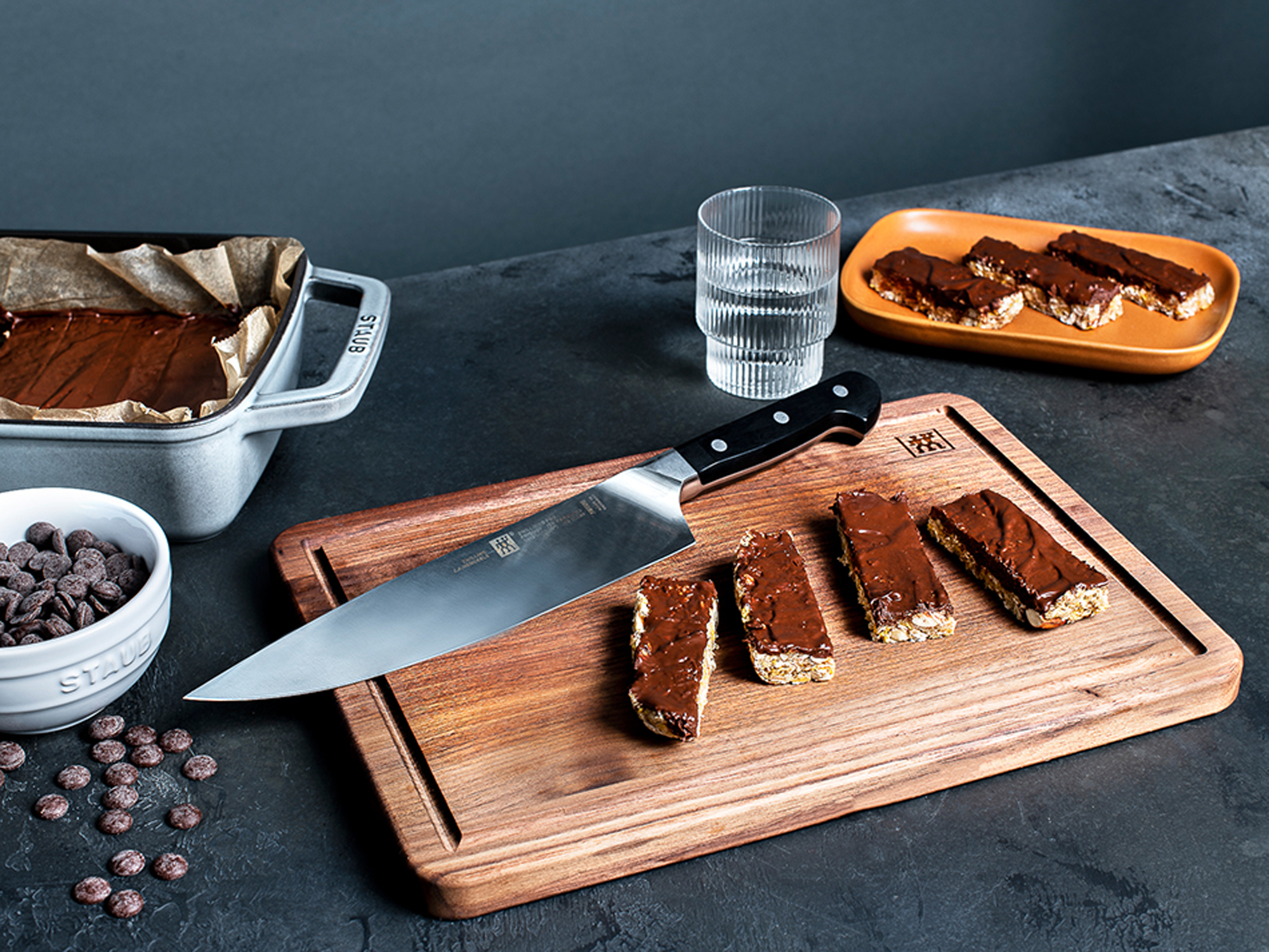 Deska do krojenia z batonami oblane czekoladą, nożem i składnikami. Mroczny, nastrojowy nastrój.