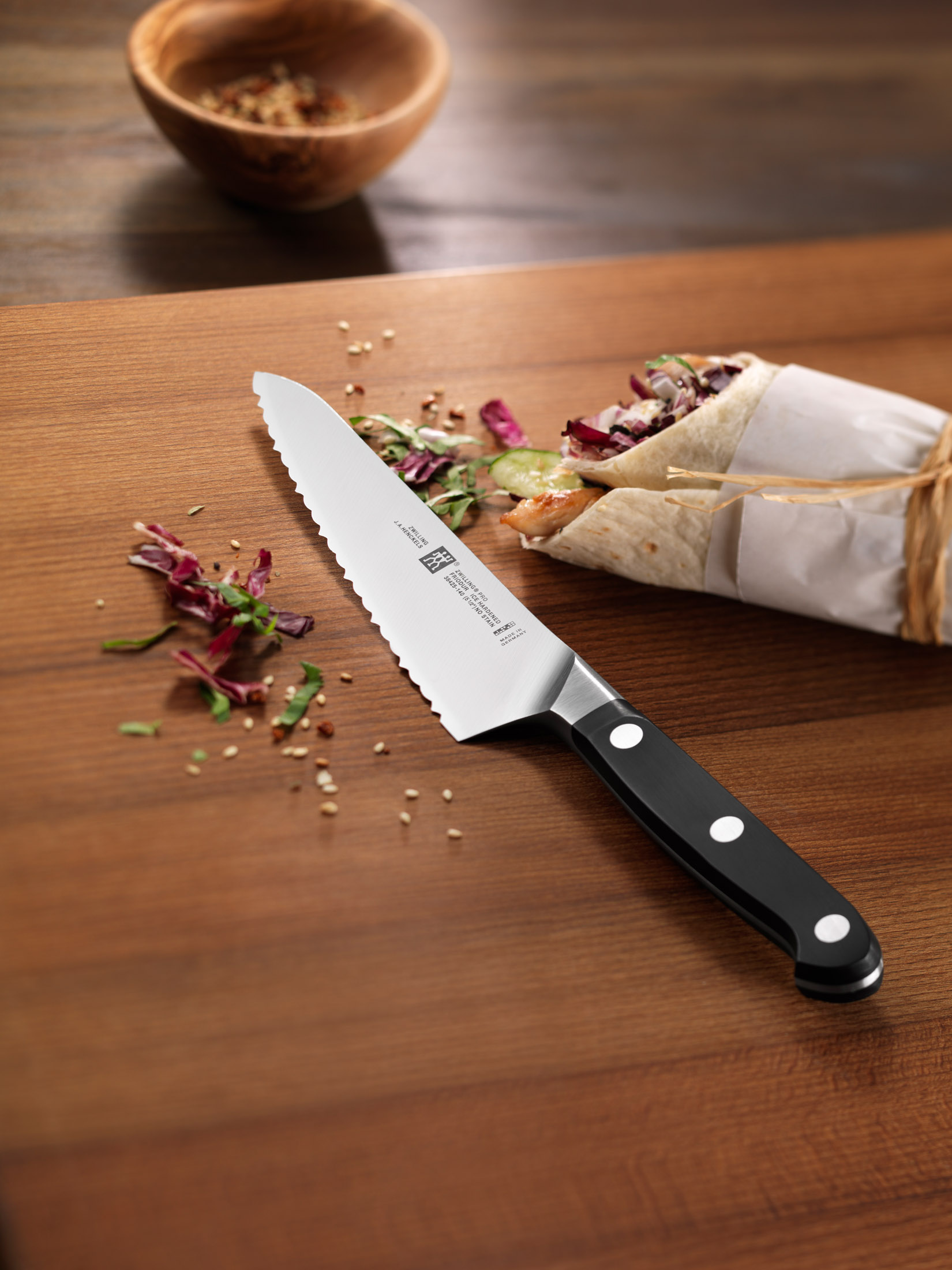 Ząbkowany nóż na drewnie, obok wrapa i miski. Sałatka i przyprawy rozrzucone.
