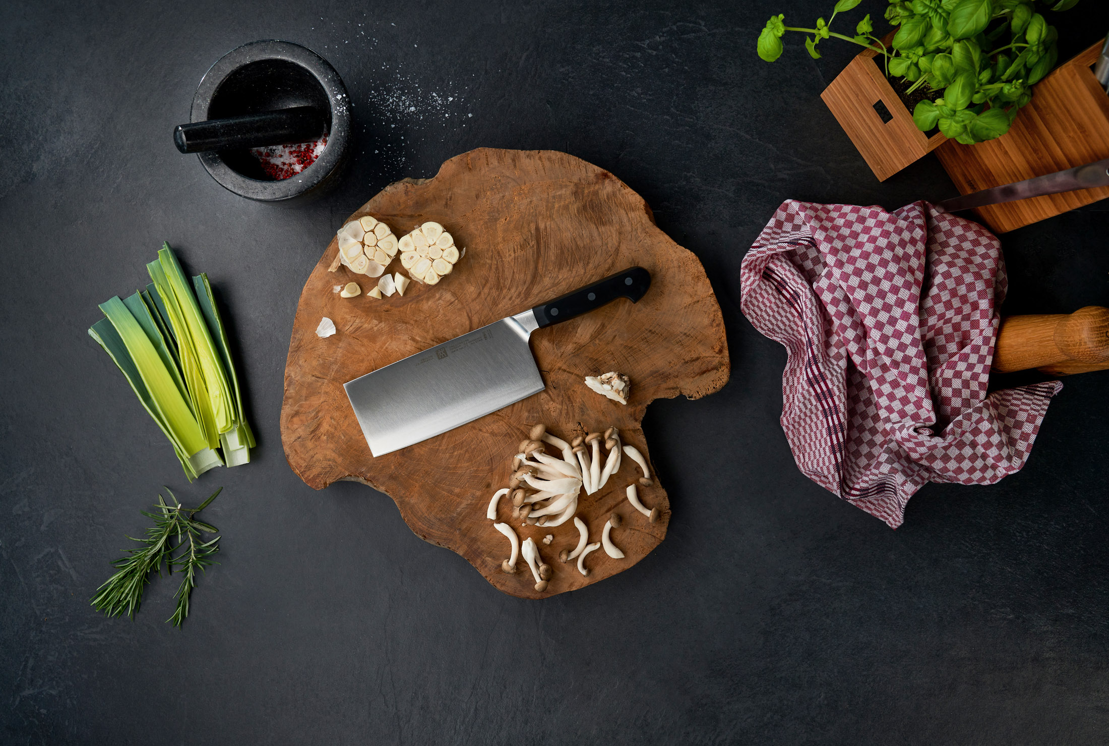 Draufsicht einer Küchenszene mit Zutaten und Werkzeugen: Lauch, Knoblauch, Pilze, Kräuter, ein Hackmesser und ein Mörser.