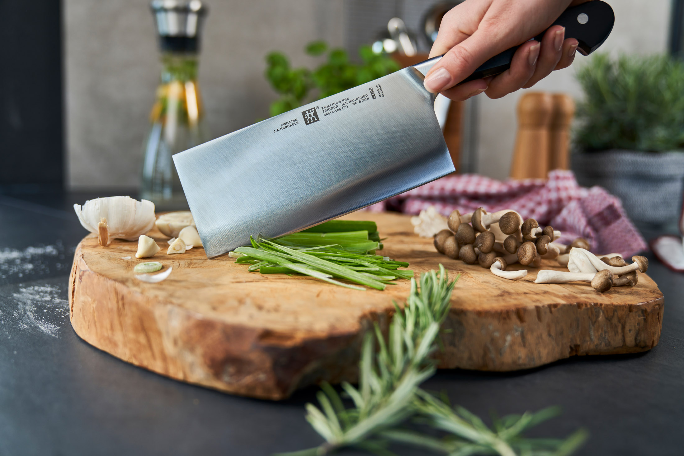 Eine Hand hält ein großes Messer über einem Holzbrett mit Knoblauch, Frühlingszwiebeln und Pilzen. Rosmarin ist im Vordergrund.