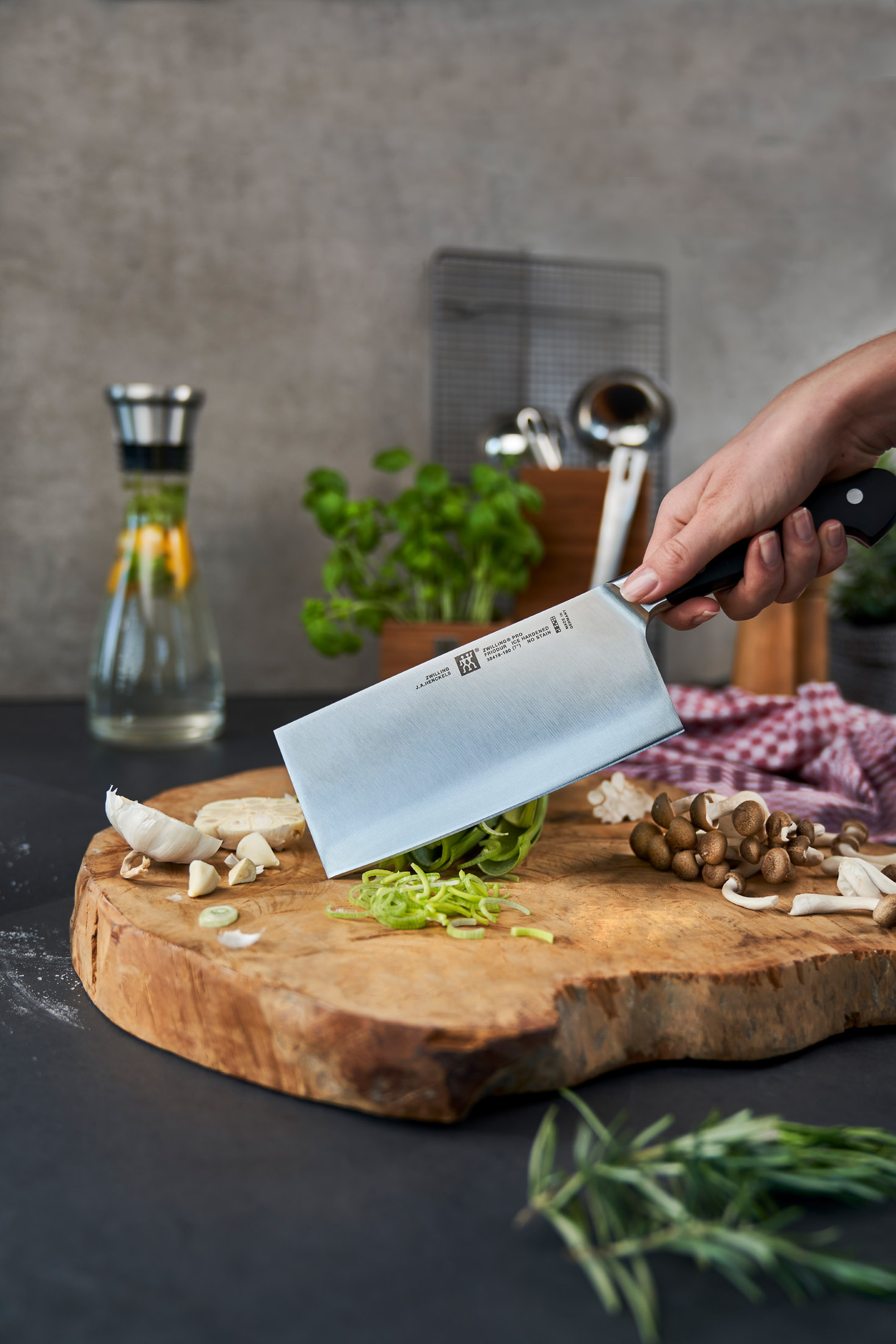 Eine Hand hält ein Hackmesser über einem Holzbrett mit Knoblauch, Pilzen und Zucchin-Nudeln. Eine Flasche und Kräuter sind im Hintergrund.