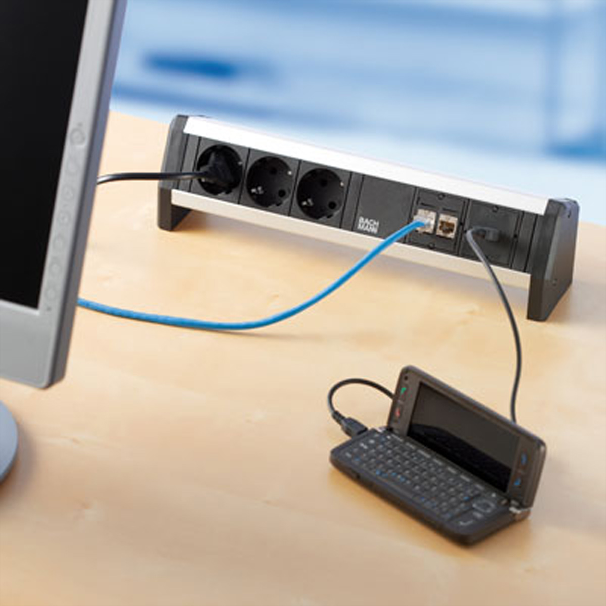 Biurko z listwą zasilającą, telefonem i monitorem. Kable łączą urządzenia. Praktyczne ustawienie biurowe.