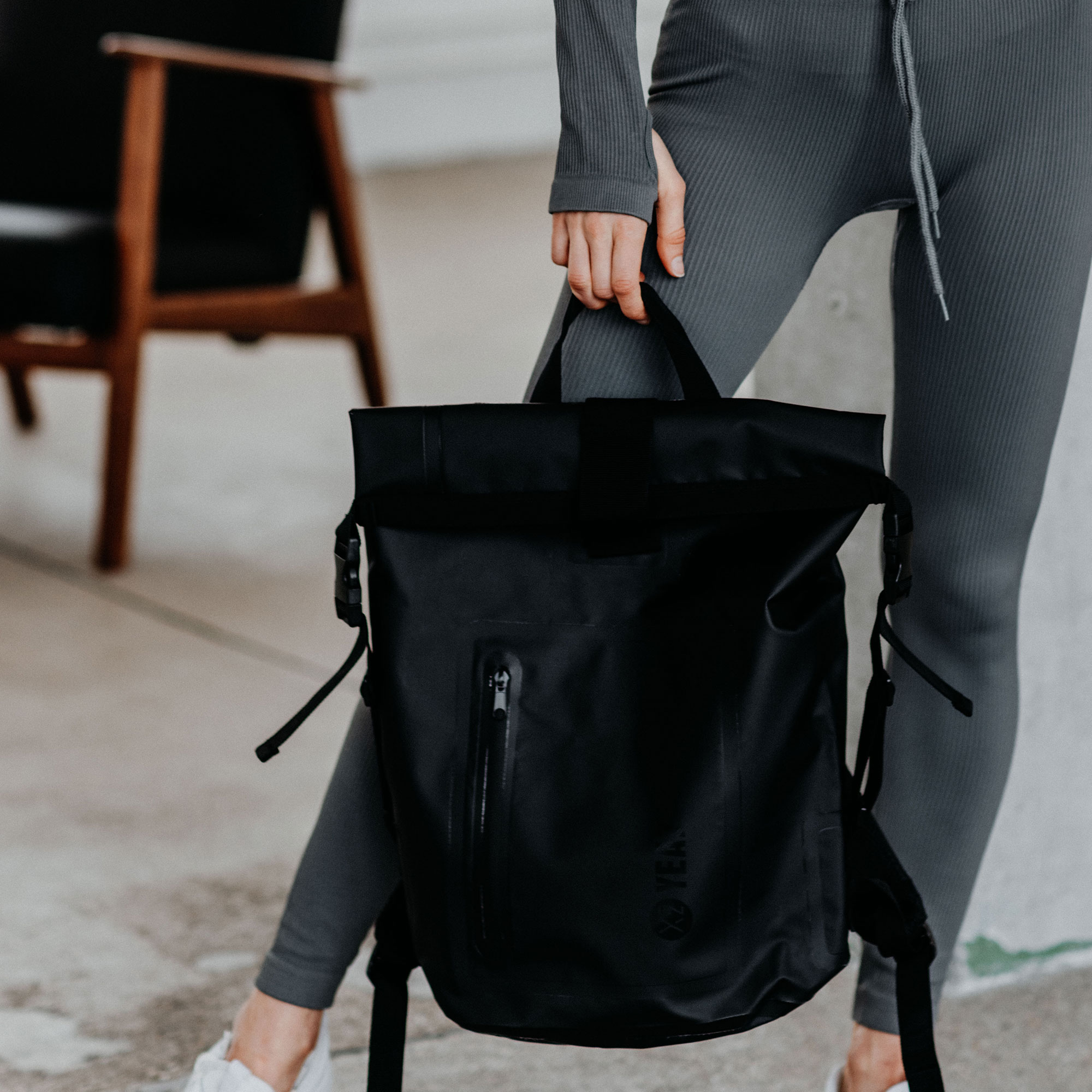 Person hält einen schwarzen Rucksack mit Rolltop-Verschluss, trägt graue Sportkleidung.
