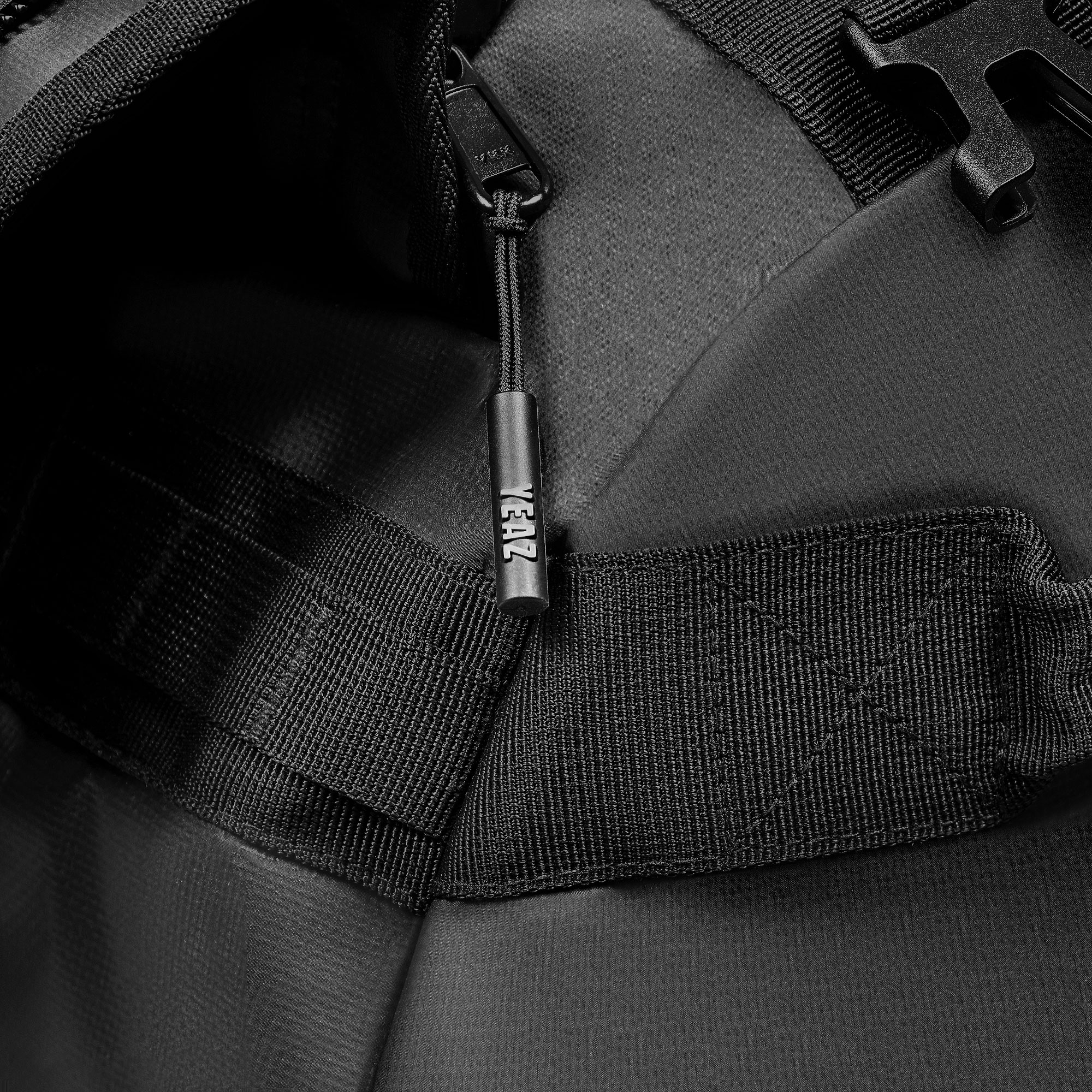 Nahaufnahme eines schwarzen Rucksacks mit Gurten und einem Reißverschlussanhänger mit dem Text 'YEAZ'.