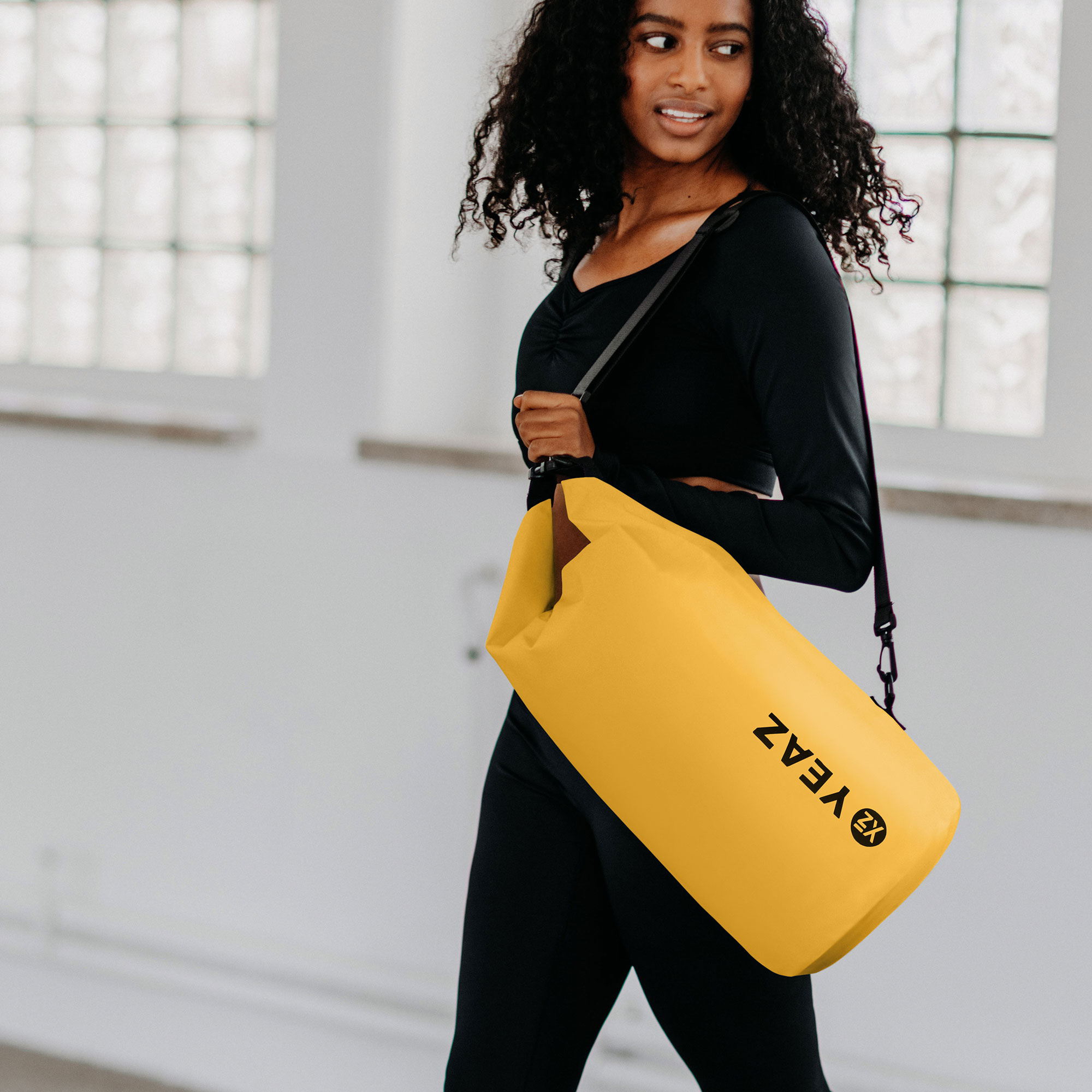 Una mujer con ropa negra lleva una bolsa amarilla sobre el hombro. Fondo blanco.