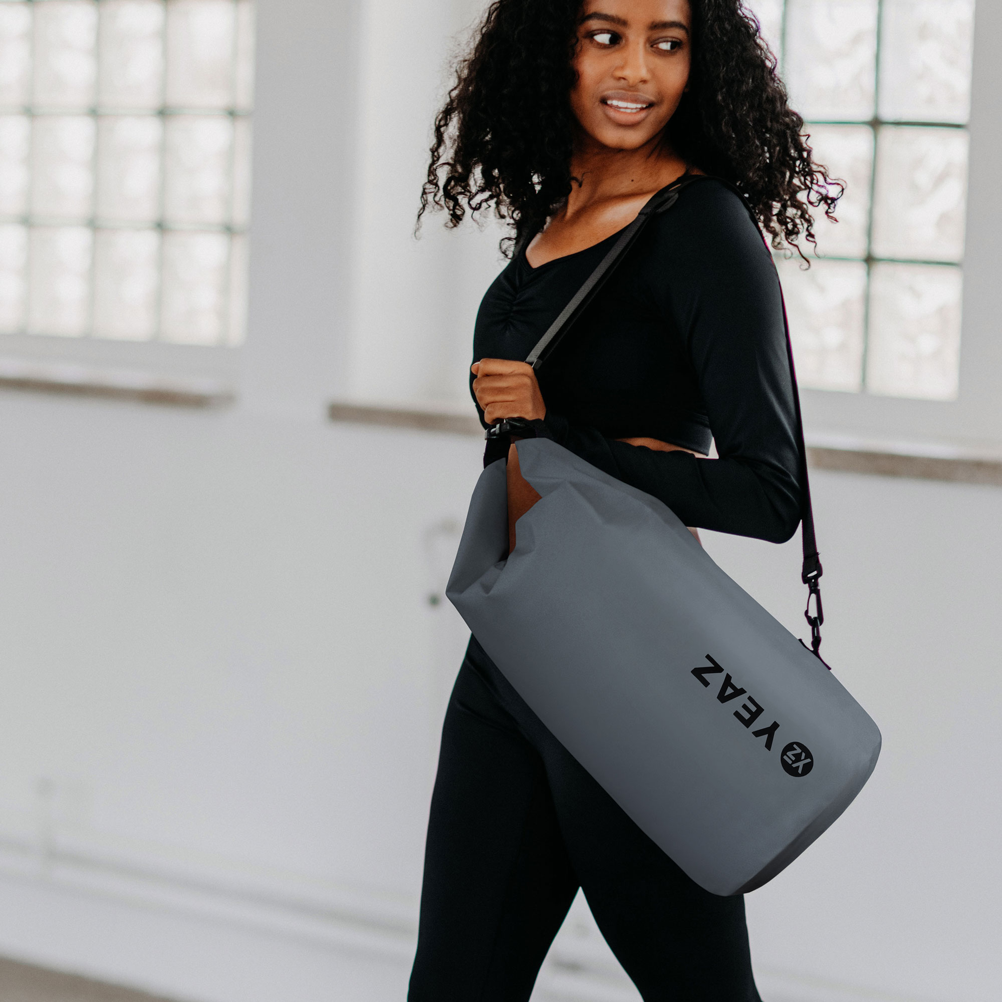 Una mujer que lleva una bolsa de gimnasia gris, caminando en un espacio brillante. Viste ropa deportiva negra.
