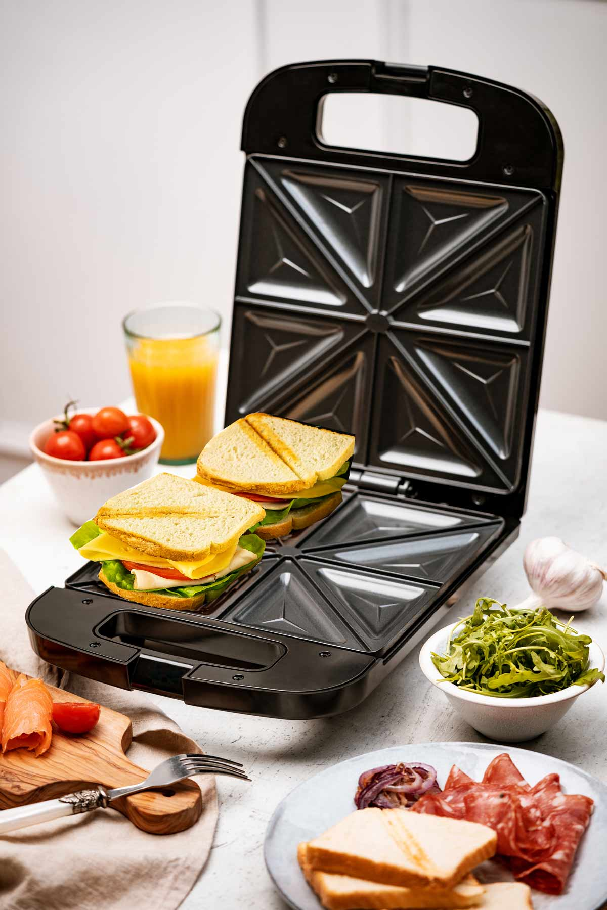 Opiekacz do kanapek jest otwarty, z dwoma kanapkami w środku. Widoczne są składniki, takie jak pomidory, ser i rukola.