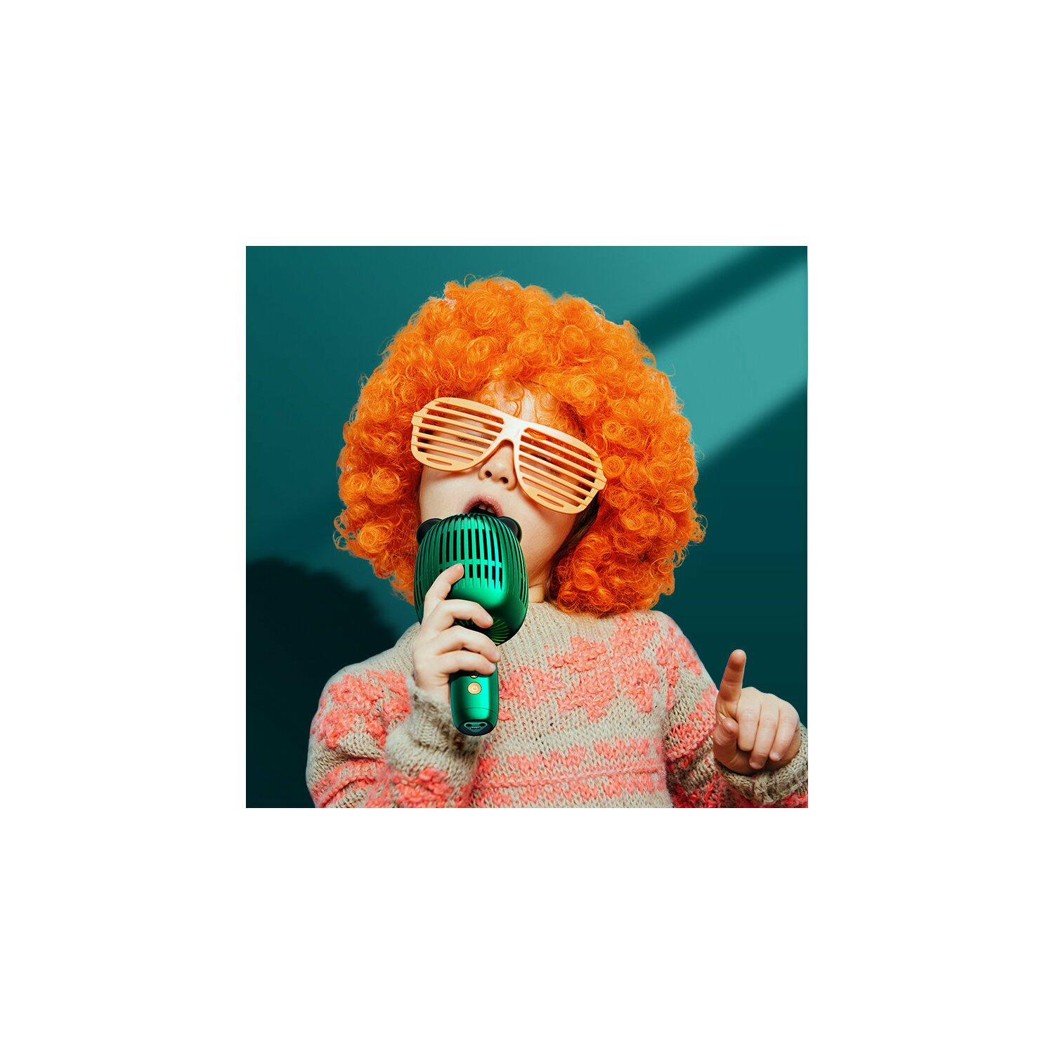 Kind mit orangefarbener Perücke und Brille singt in ein grünes Mikrofon. Zeigt mit dem Finger nach oben, grüner Hintergrund.