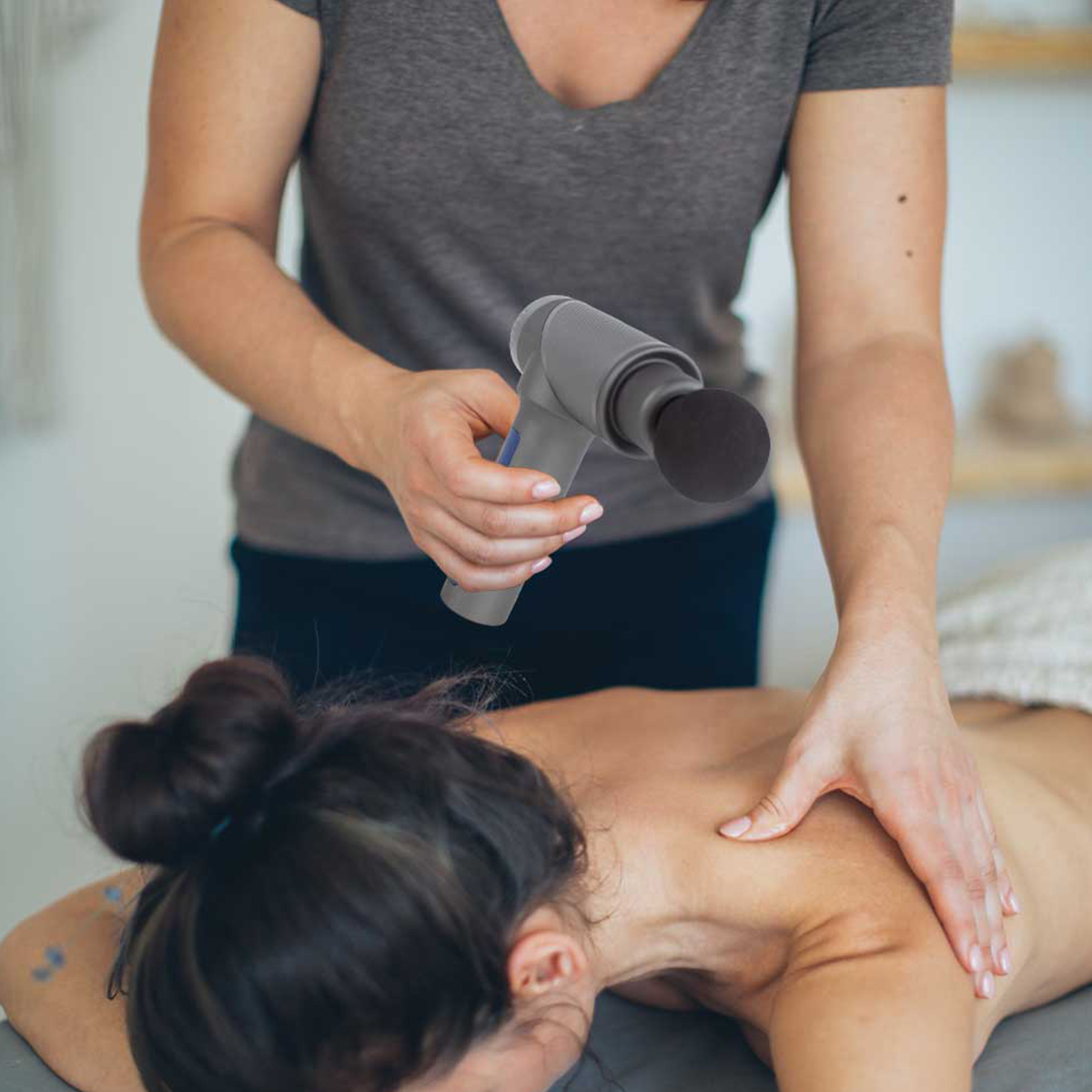 Osoba wykonująca masaż za pomocą szarego pistoletu do masażu na plecach osoby.