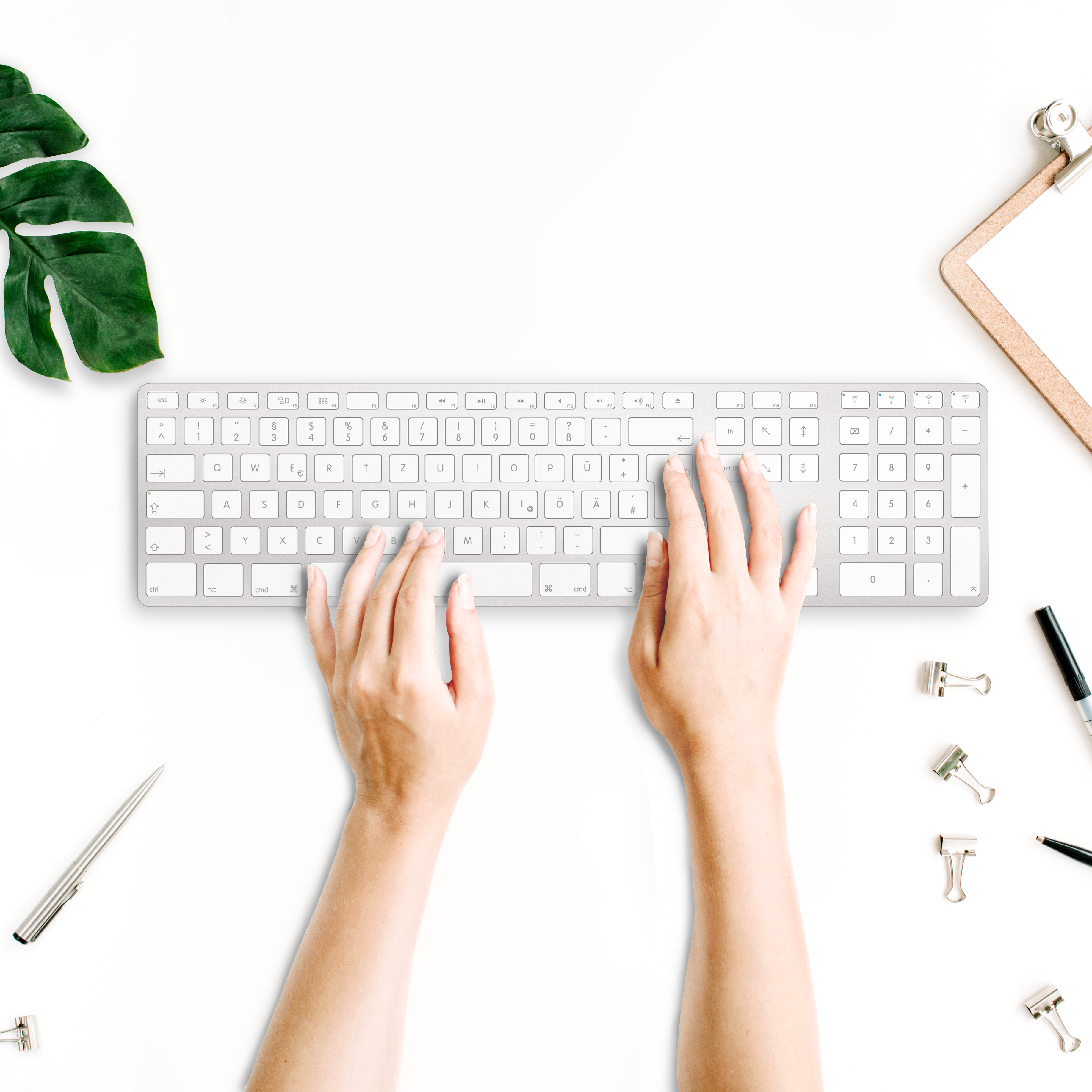 Hände, die auf einer silbernen Tastatur auf einem weißen Schreibtisch mit Büromaterial und einer Pflanze tippen.
