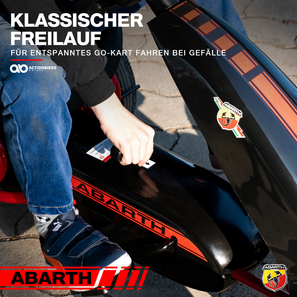 Ein schwarzes Go-Kart mit roten Streifen. Die Hand eines Kindes ist darauf platziert. Die Marke 'ABARTH' ist sichtbar.