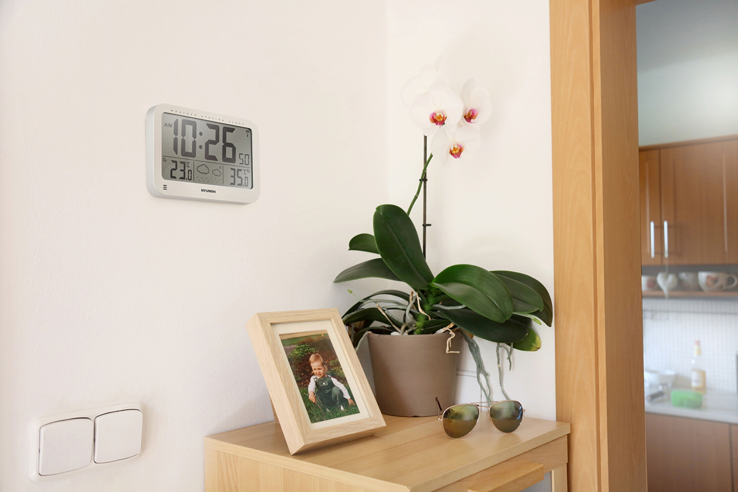 Eine Digitaluhr, ein Fotorahmen und eine Orchidee stehen auf einem Holztisch.