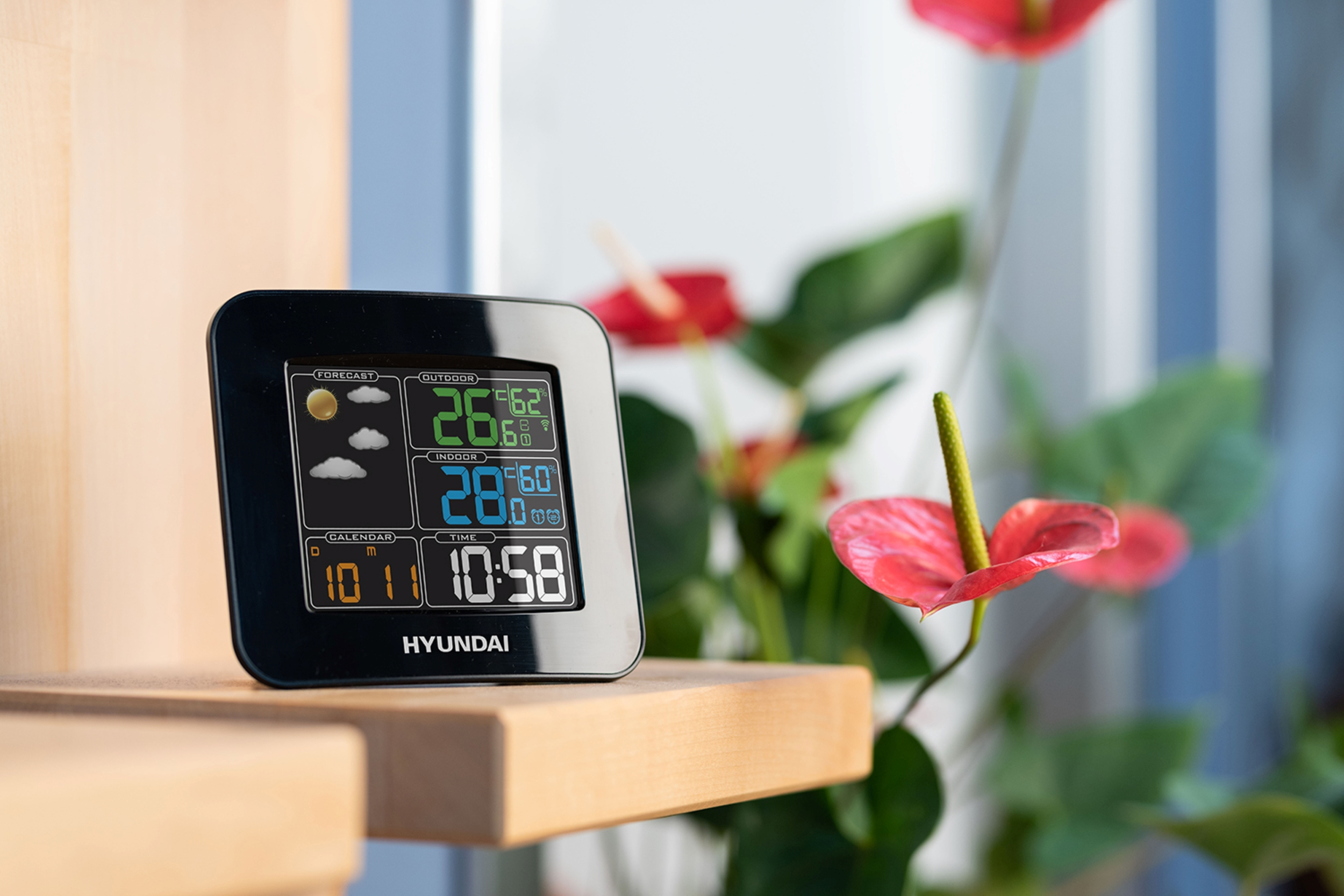 Schwarze Wetterstation mit Anzeige, neben roten Blumen und grünen Blättern.
