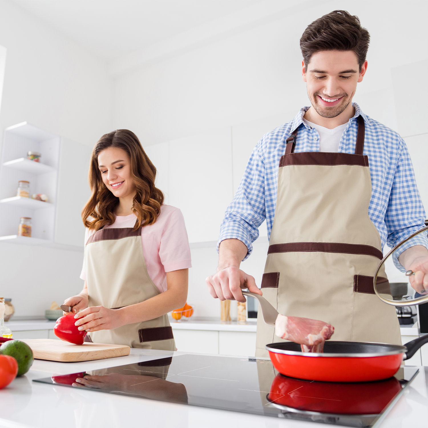 Una pareja cocinando en una cocina moderna; uno voltea comida en una sartén roja.