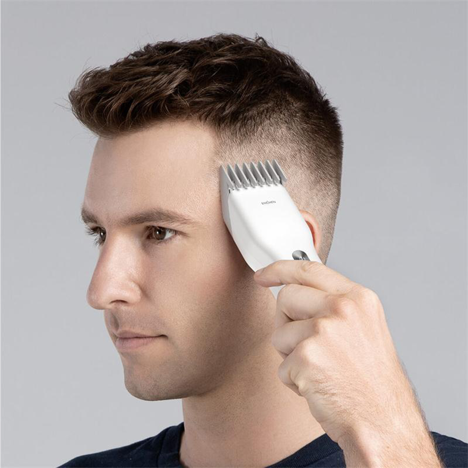 Mann, der sich mit einem weißen Haarschneider die Haare schneiden lässt, Seitenansicht, hellgrauer Hintergrund.