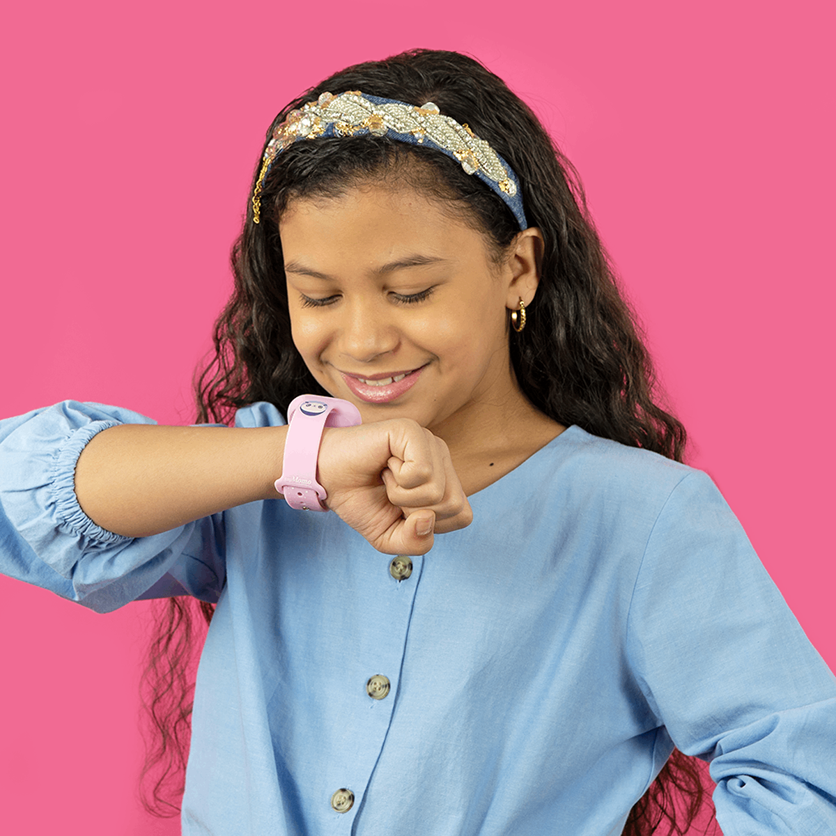 Una niña con camisa azul mira un smartwatch rosa en su muñeca contra un fondo rosa.