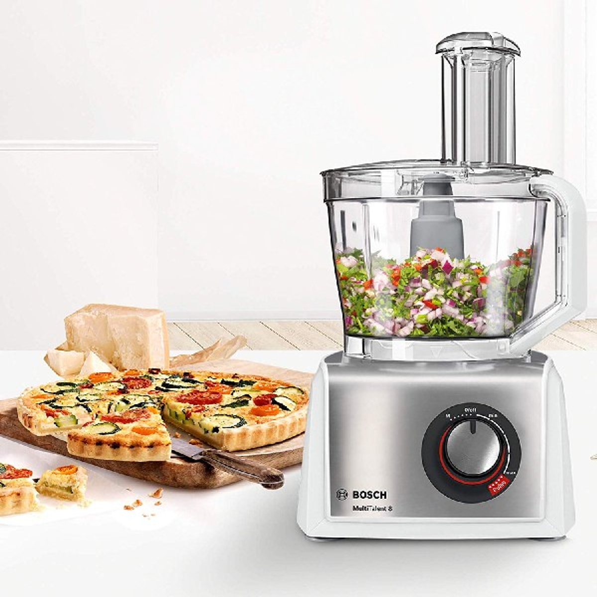 Procesador de alimentos con ingredientes y comida preparada sobre superficie blanca.