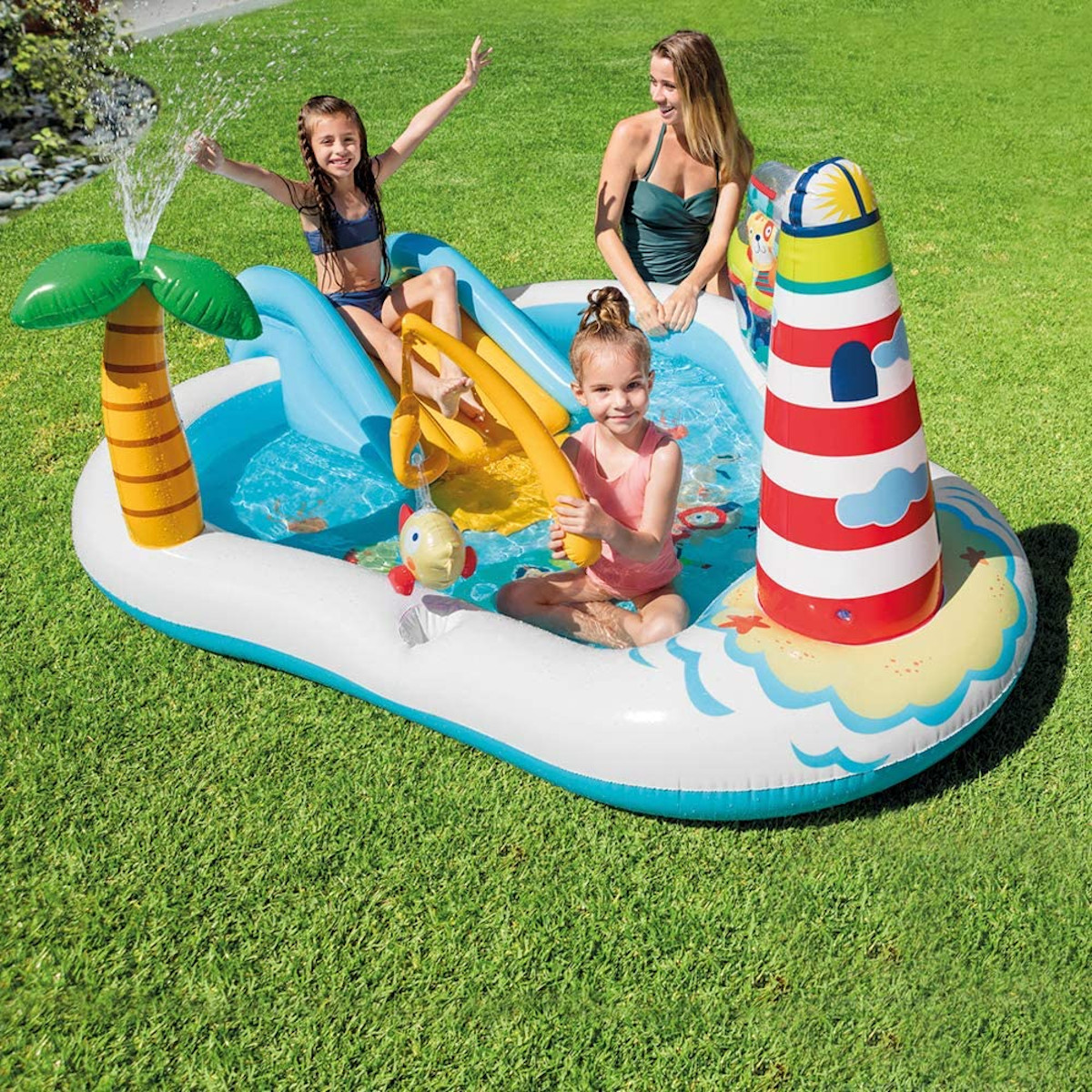 Bambini giocano in una piscina gonfiabile con scivolo, palma, faro e donna sorridente.
