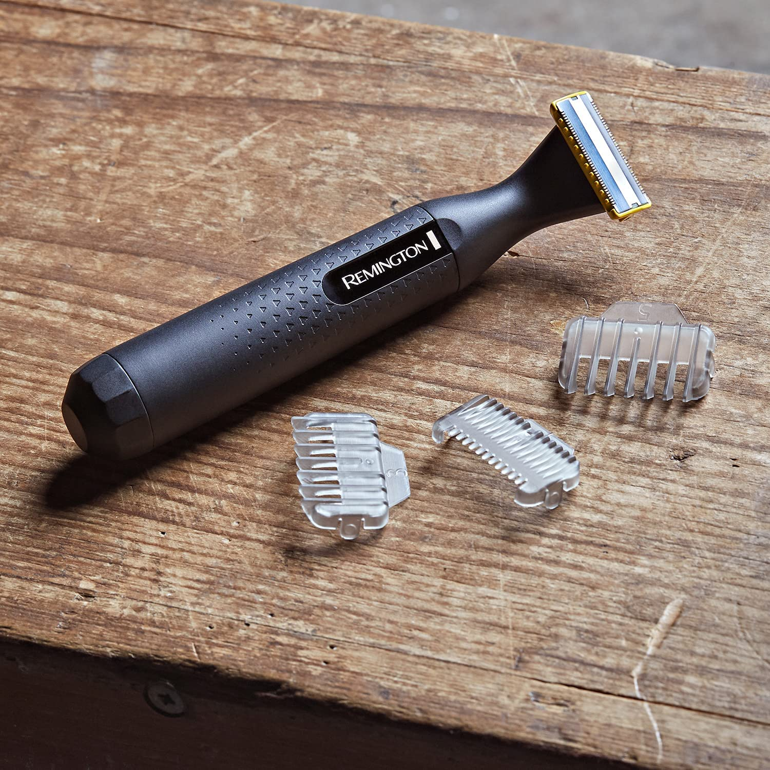 Una recortadora corporal Remington gris y tres accesorios de plástico transparente, sobre una superficie de madera.
