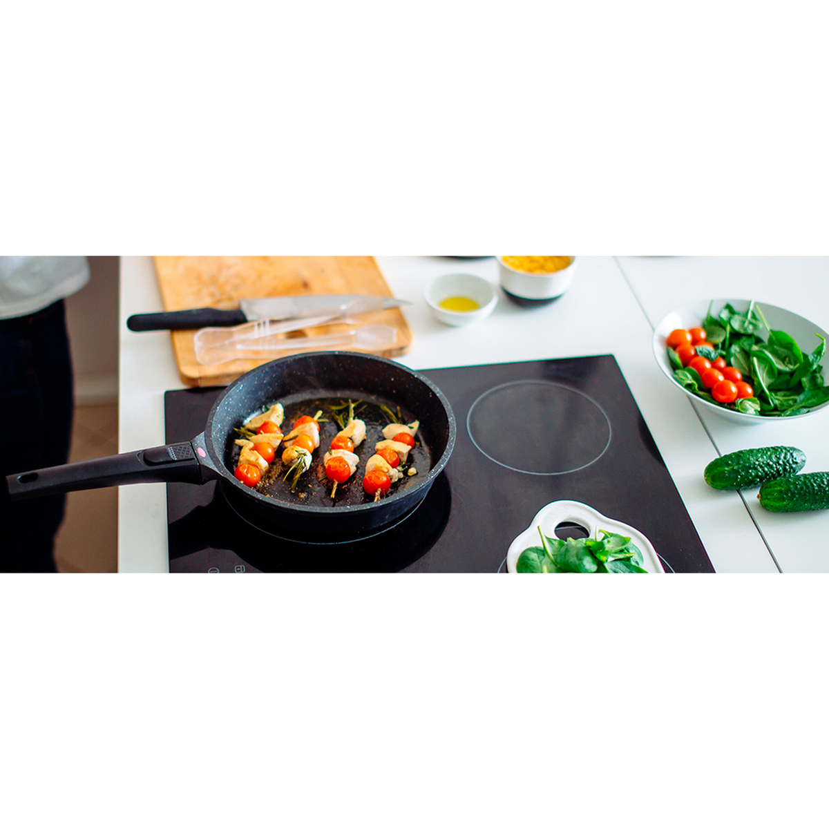 Comida cocinándose en una placa de inducción negra, con tomates, pepinos y verduras cerca.