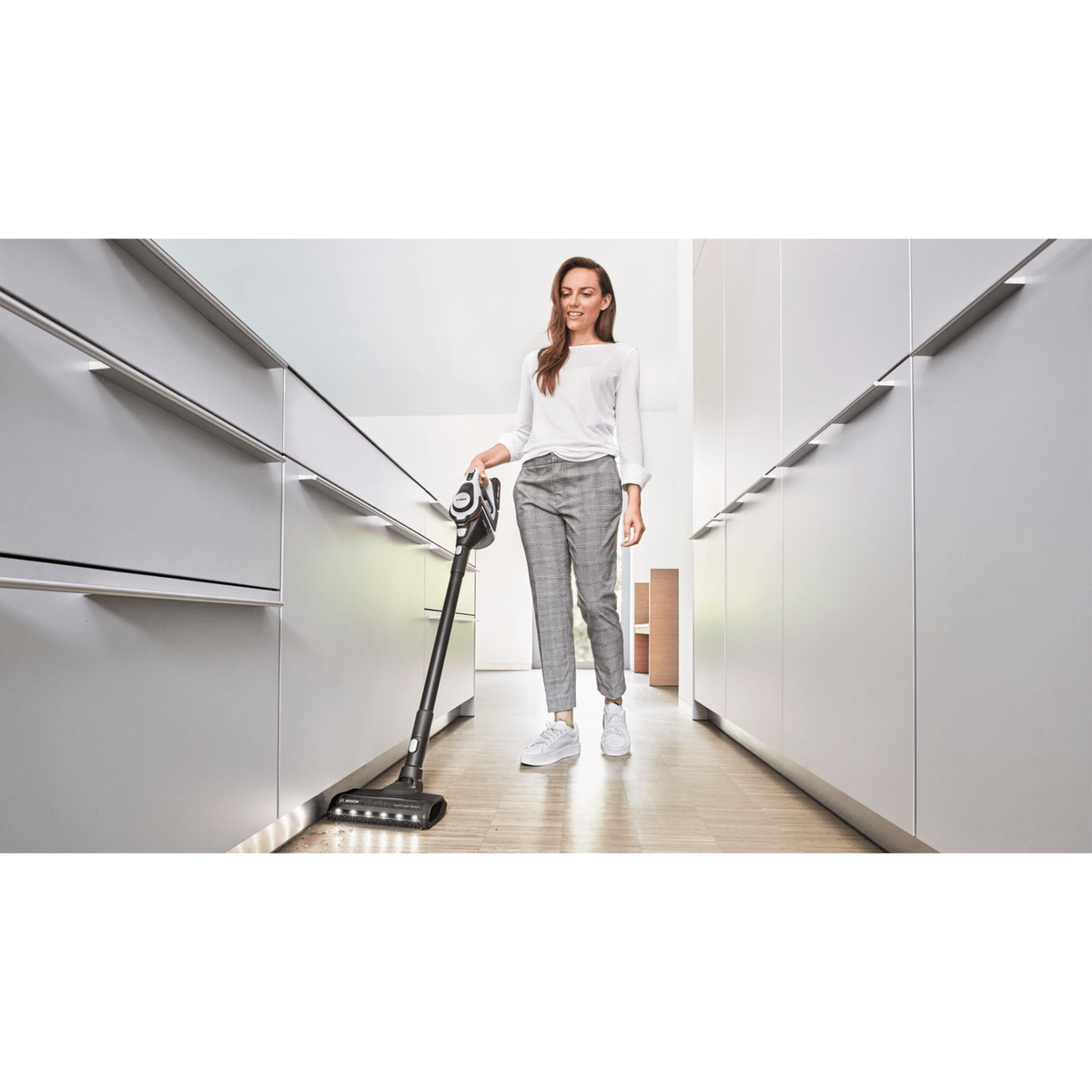 Mujer aspirando con una aspiradora en blanco y negro. Lleva camisa blanca y pantalones grises.