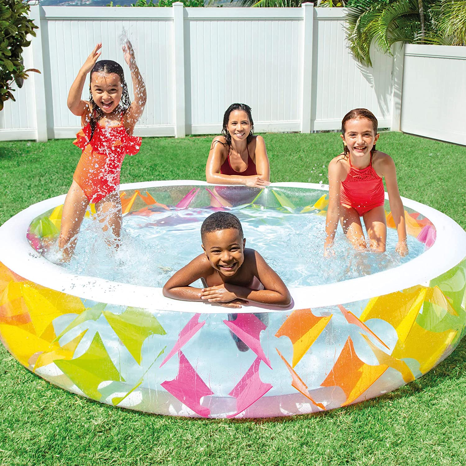 Kinder spielen in einem bunten aufblasbaren Pool auf Gras, im Hintergrund ein weißer Zaun.