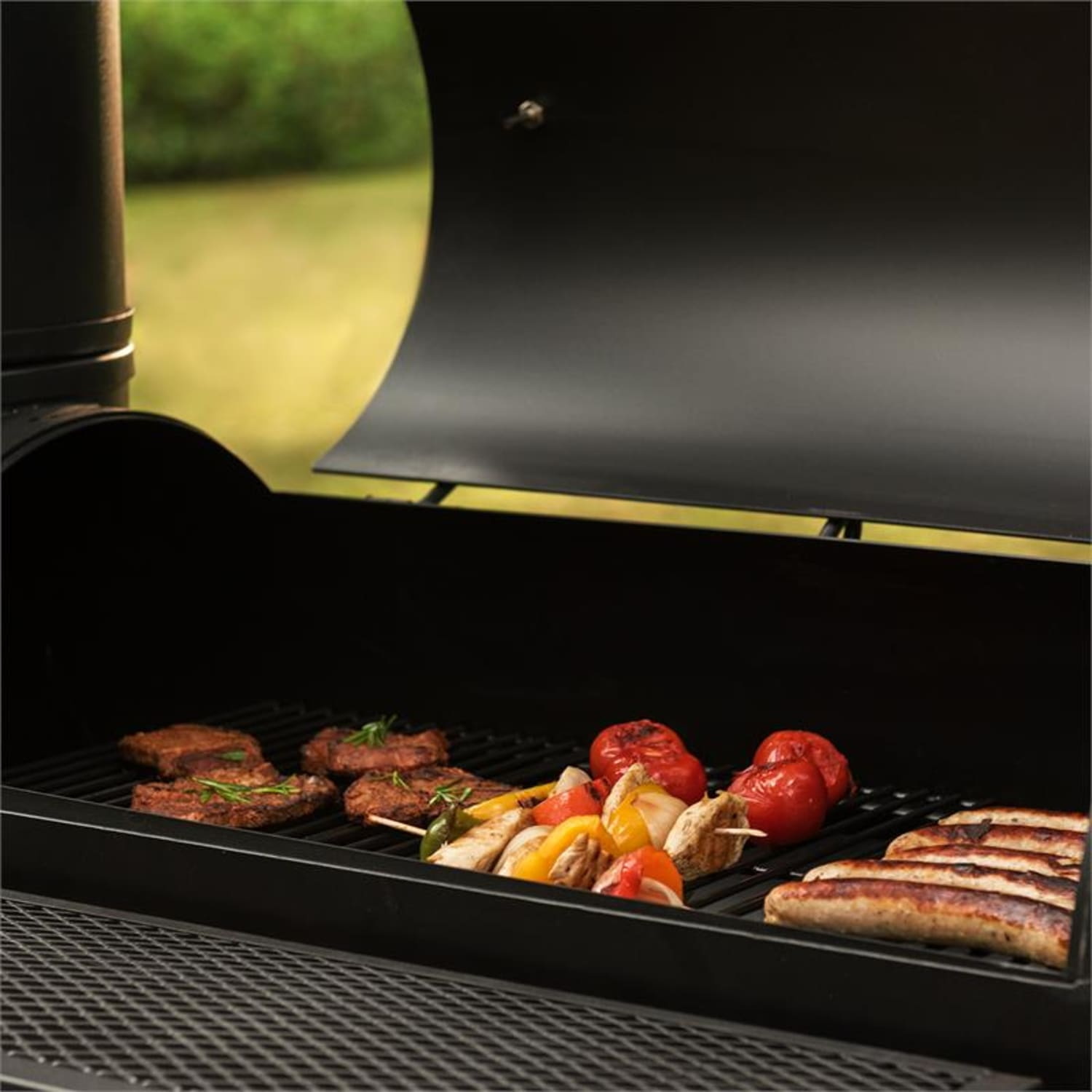 Nahaufnahme eines Grills mit Fleisch, Gemüse und Würstchen. Der Deckel ist offen, das Essen sichtbar.