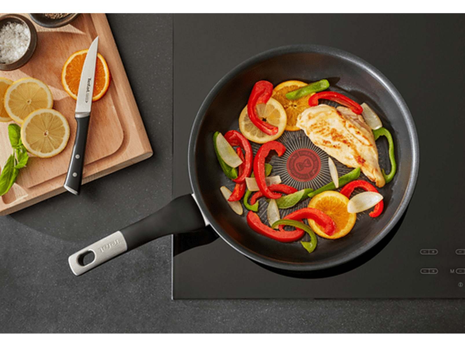 Poêle avec poulet, tranches de citron et poivrons sur plaque noire.