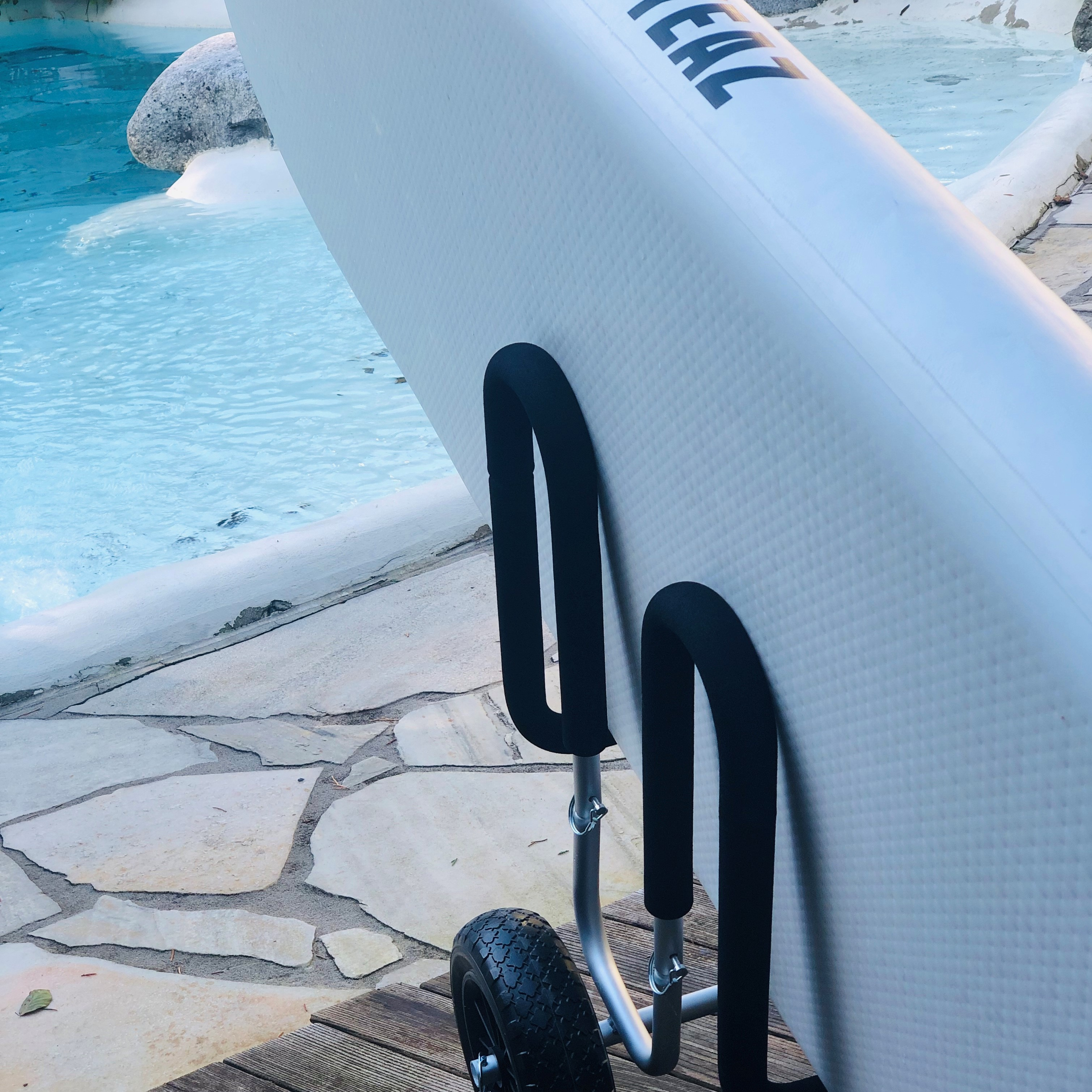 Ein weißes Paddleboard auf einem Wagen. Blaues Wasser und ein Steinweg im Hintergrund.