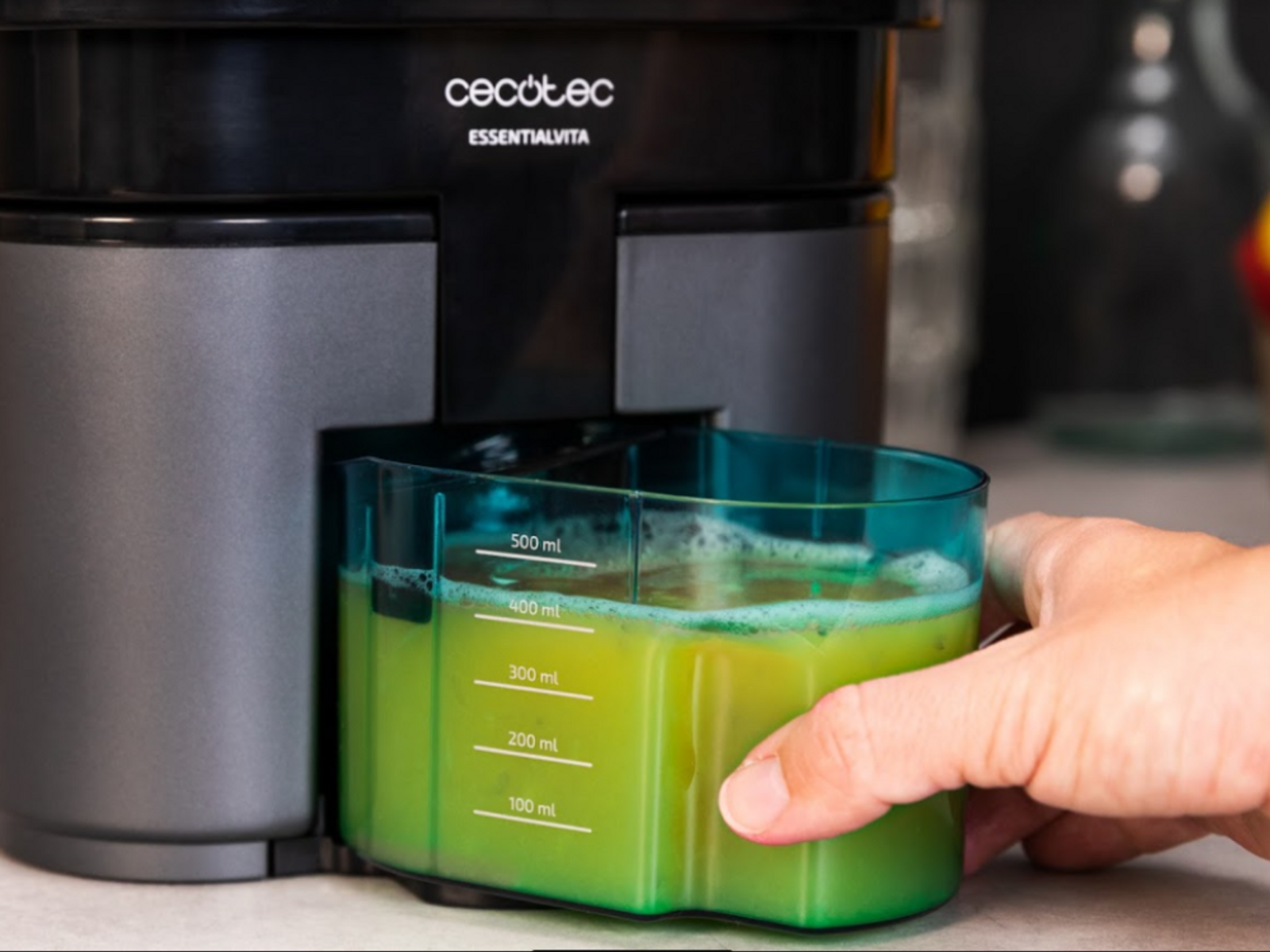 Una mano tiene un contenitore pieno di succo verde, vicino a un estrattore nero con l'etichetta 'Cecotec EssentialVita'.