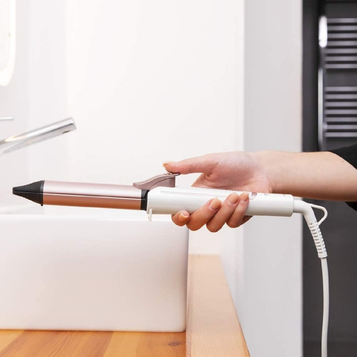 Dłoń osoby trzymająca lokówkę w kolorze różowego złota i bieli w pobliżu białej umywalki.