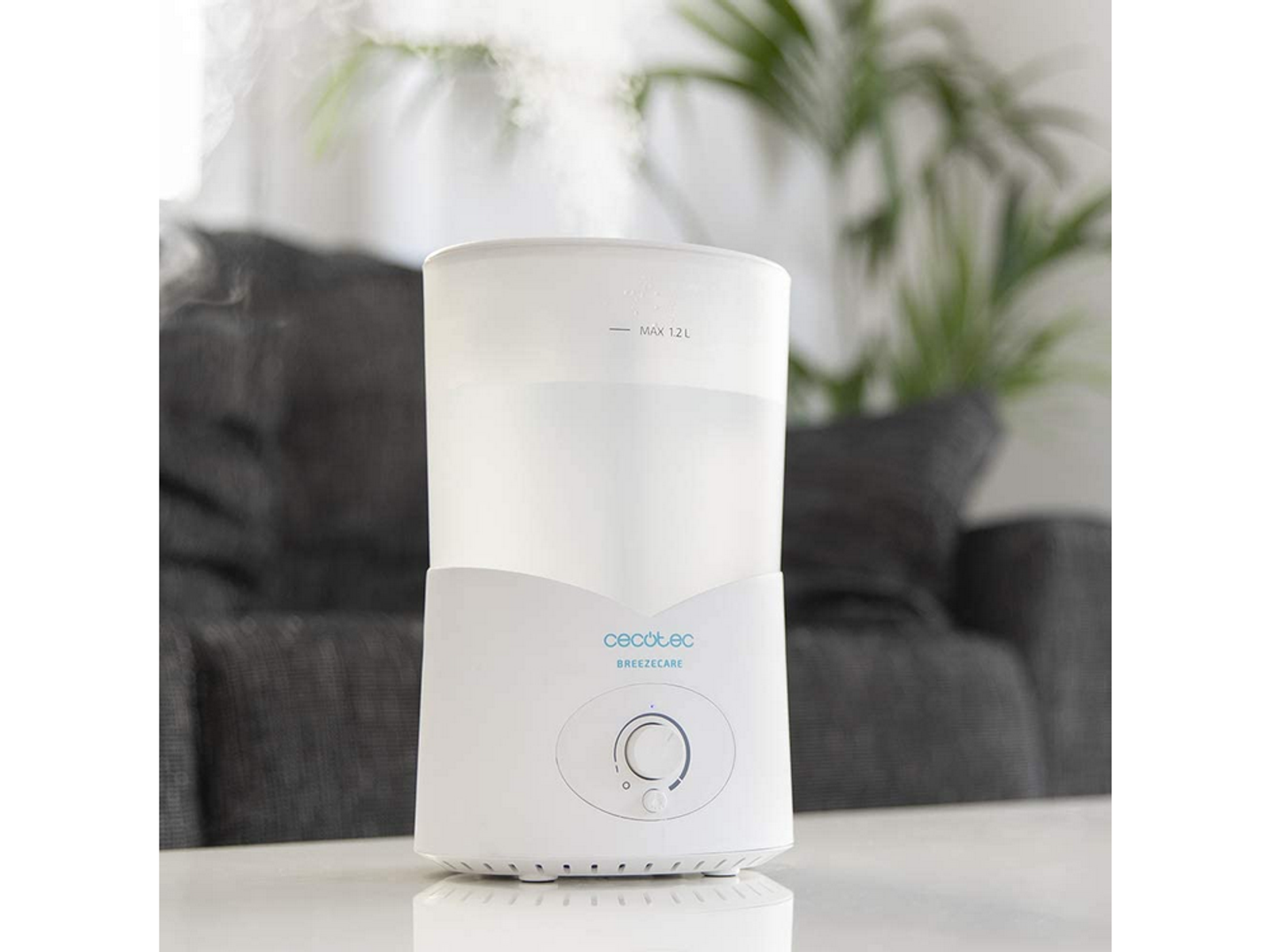 Biały stół z białym nawilżaczem i rozmytą szarą sofą z zieloną rośliną w tle.